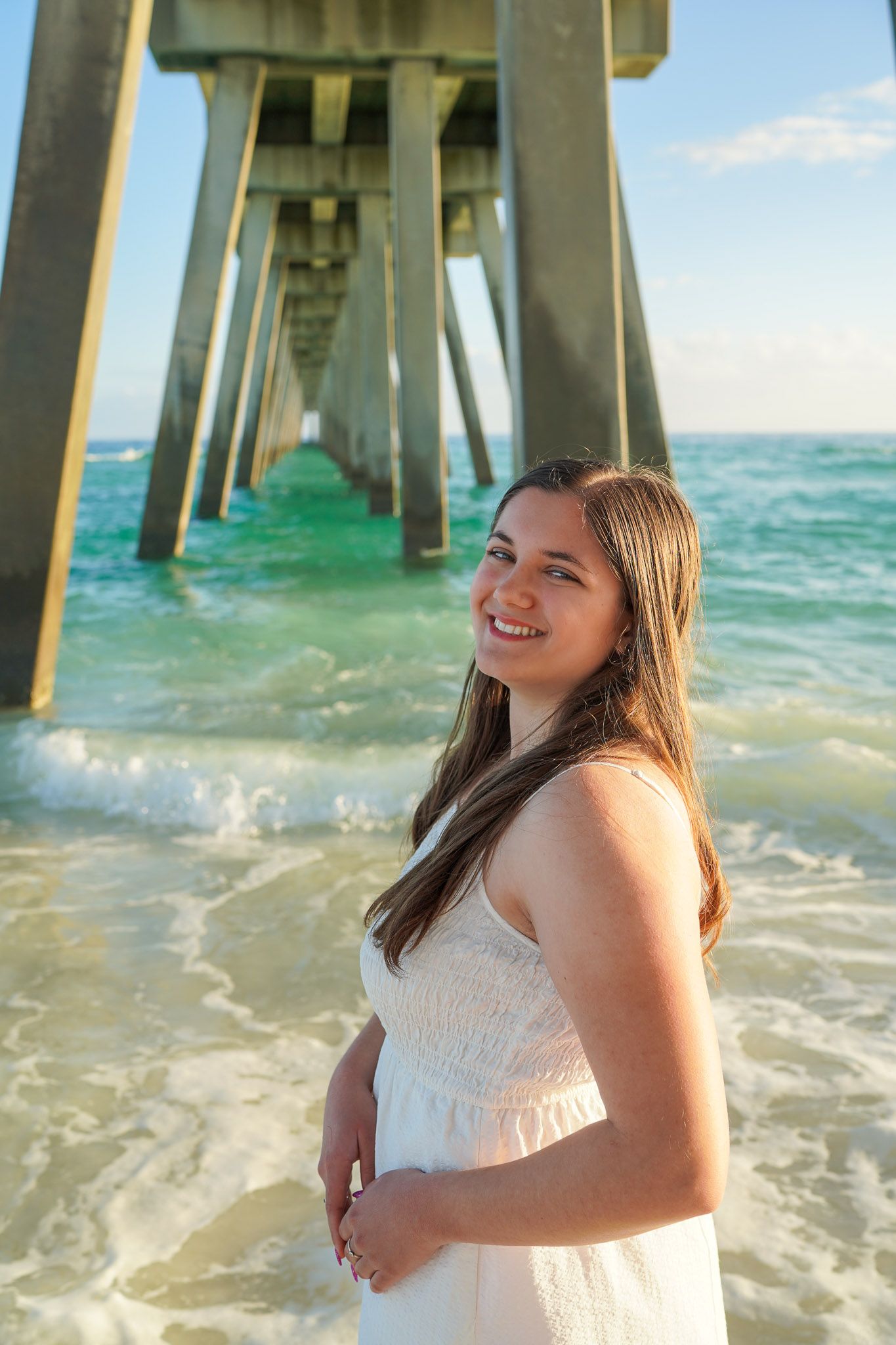 Happy young woman in white dress laughing on beach with ocean pier in background.