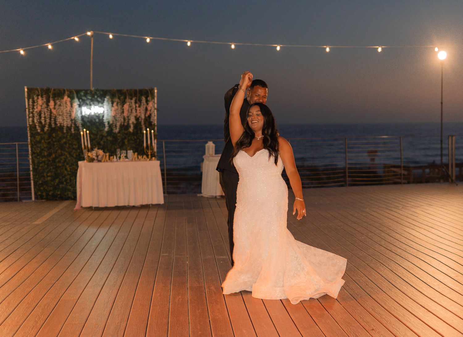 Bride and groom share their first dance on an outdoor deck at dusk with string lights overhead.