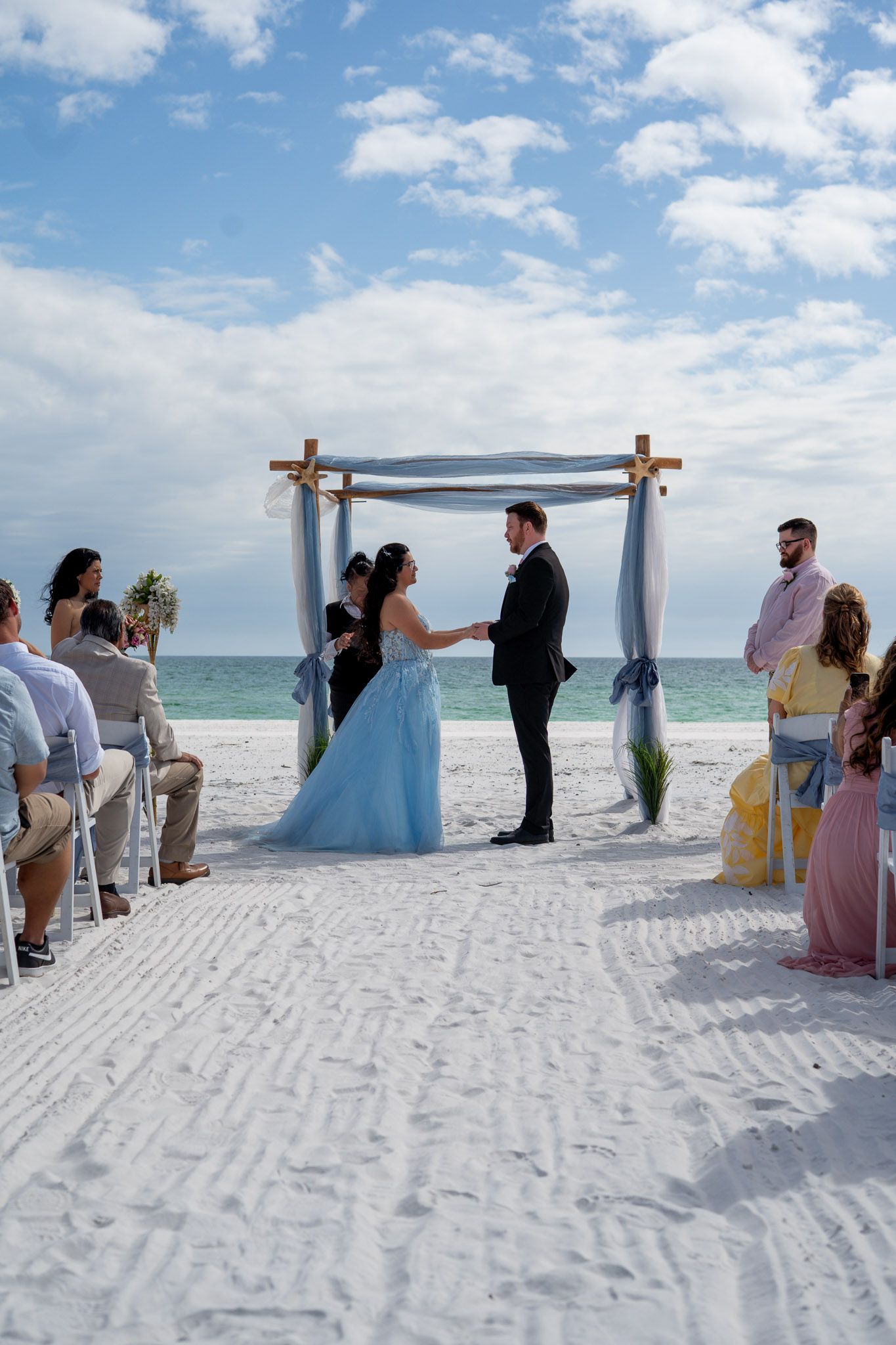 Couple exchanges vows at a beach wedding ceremony under a wooden arch draped with blue fabric, guests seated on white sand.