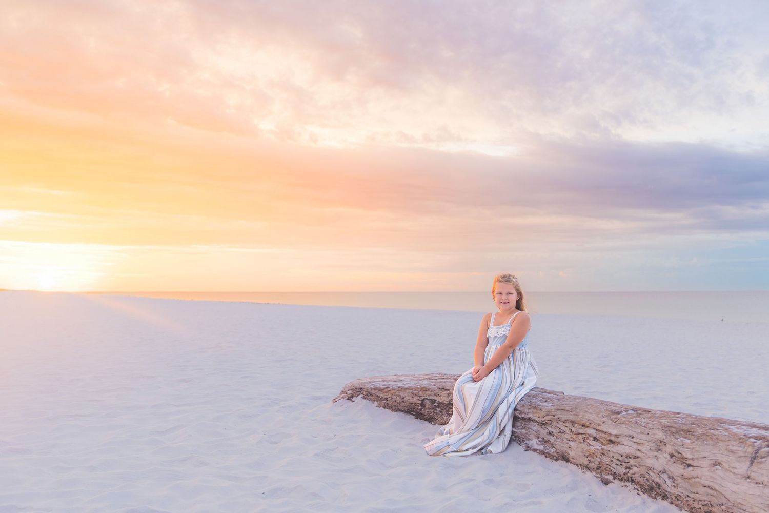 Woman in white dress sitting on driftwood at sunset on white sand beach.