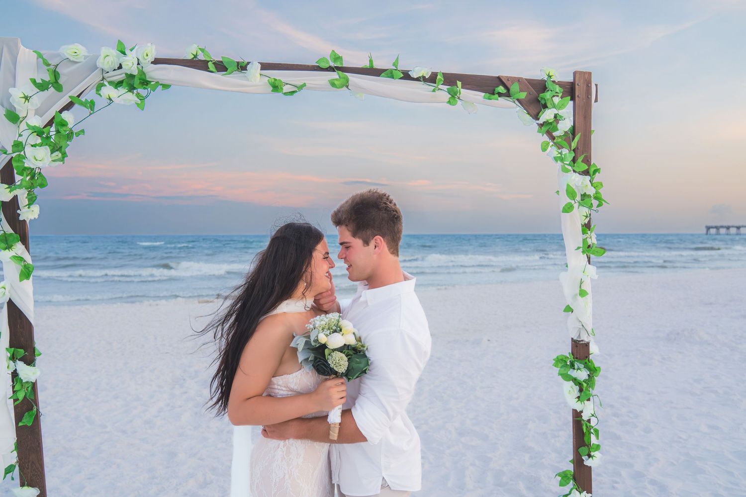 Newlyweds embrace under green ivy arch on sandy beach, bride holding white bouquet, ocean sunset in background.