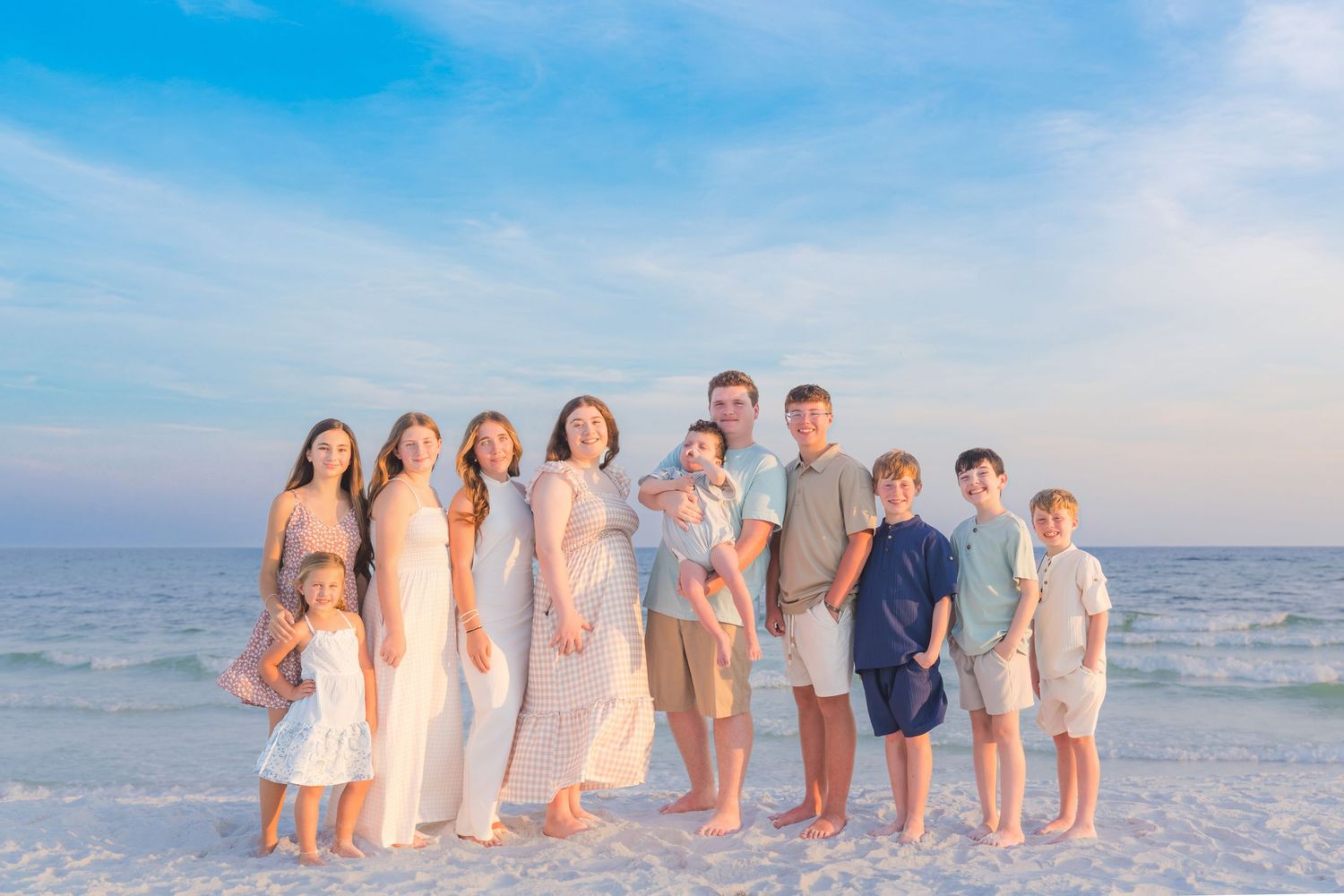 Large family posing together on a white sand beach at sunset, wearing coordinated neutral and blue outfits.