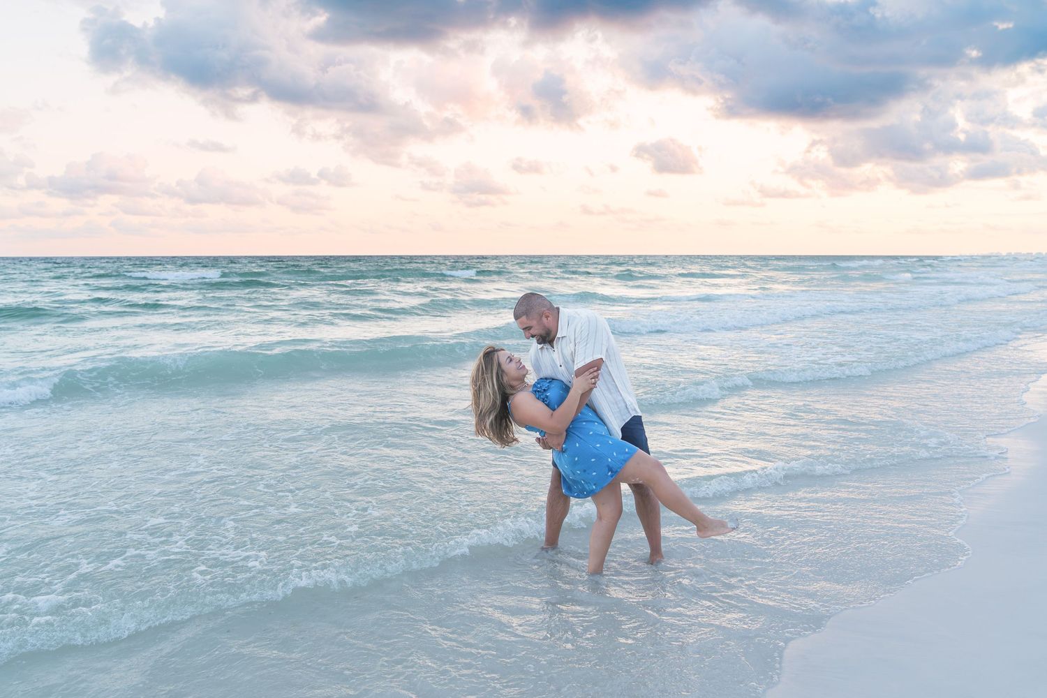 Couple dancing romantically in ocean waves at sunset on a white sand beach.