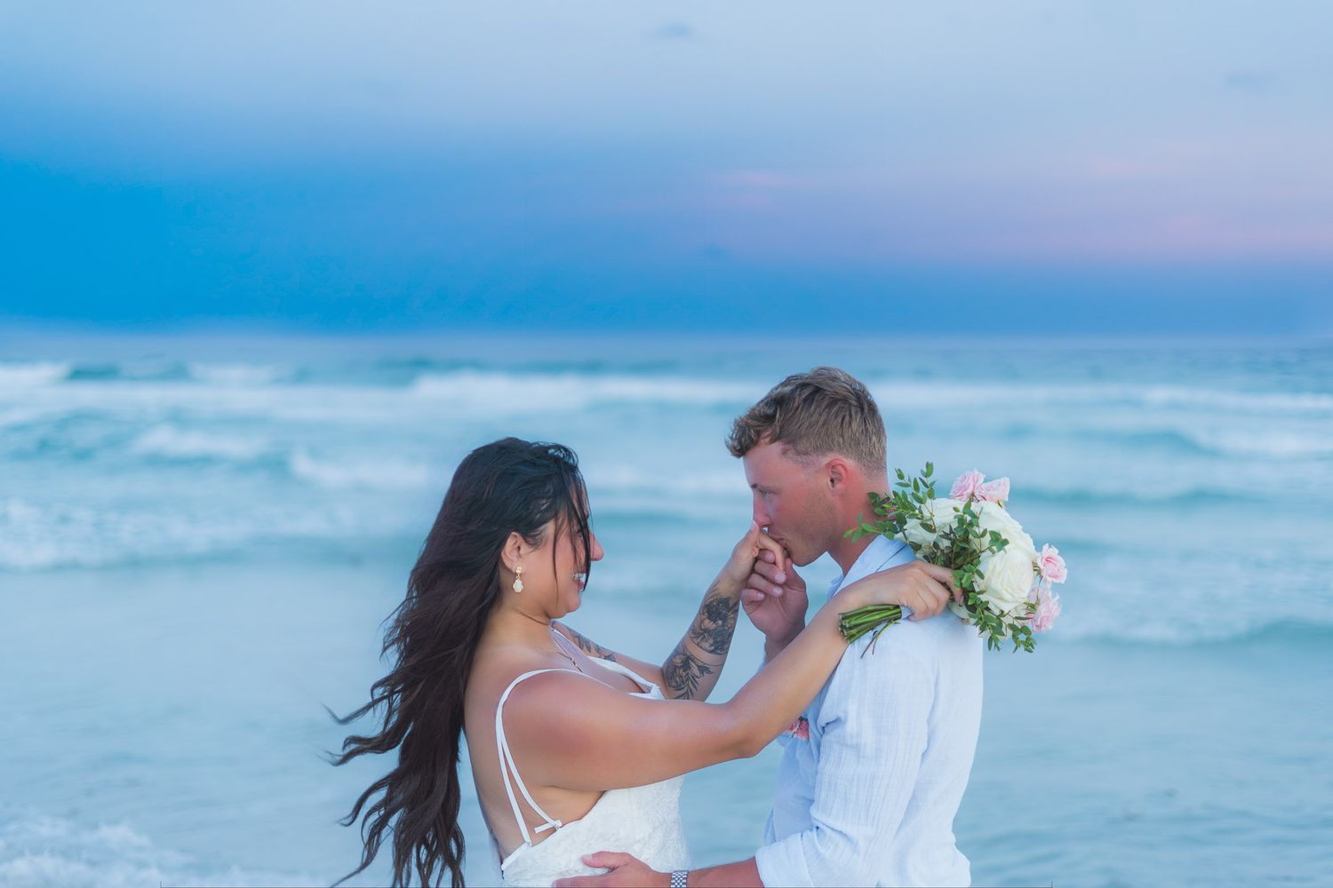 Groom kisses bride's hand on beach at dusk, bride holding pink and white bouquet with ocean waves behind them.
