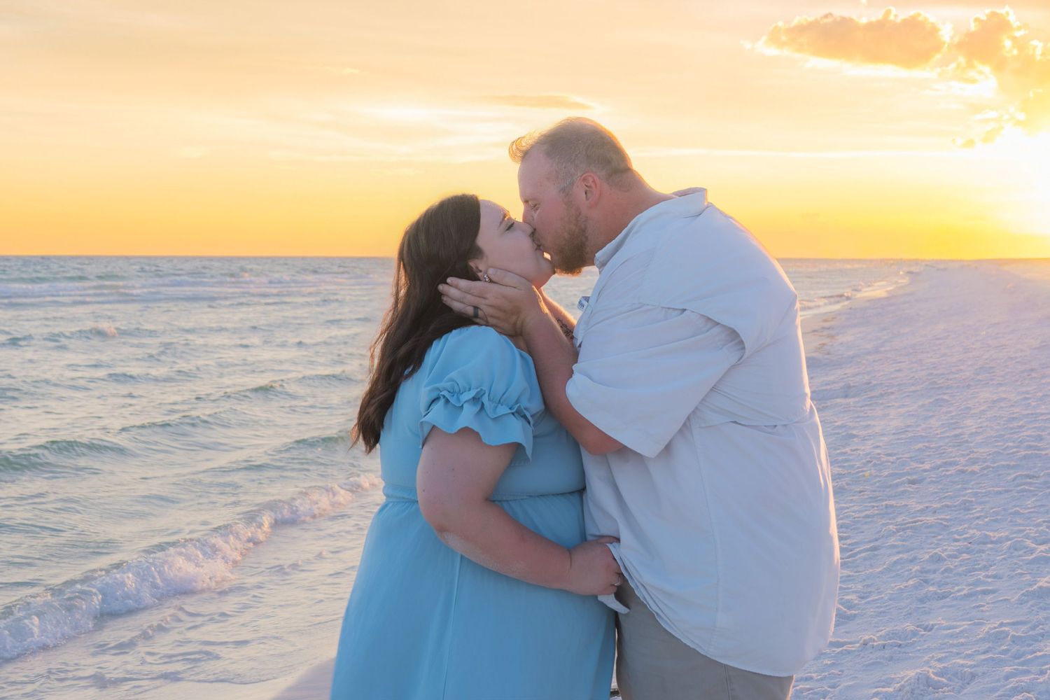Couple sharing a romantic kiss on the beach at golden sunset, woman in blue dress, waves in background.