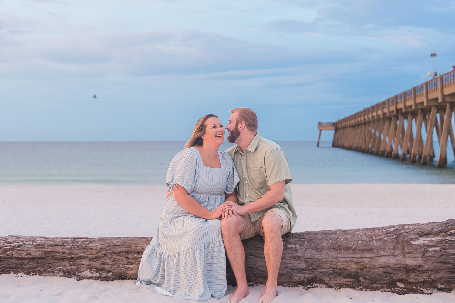 Couple sitting on driftwood log at white sand beach near pier, sharing a romantic moment at dusk.