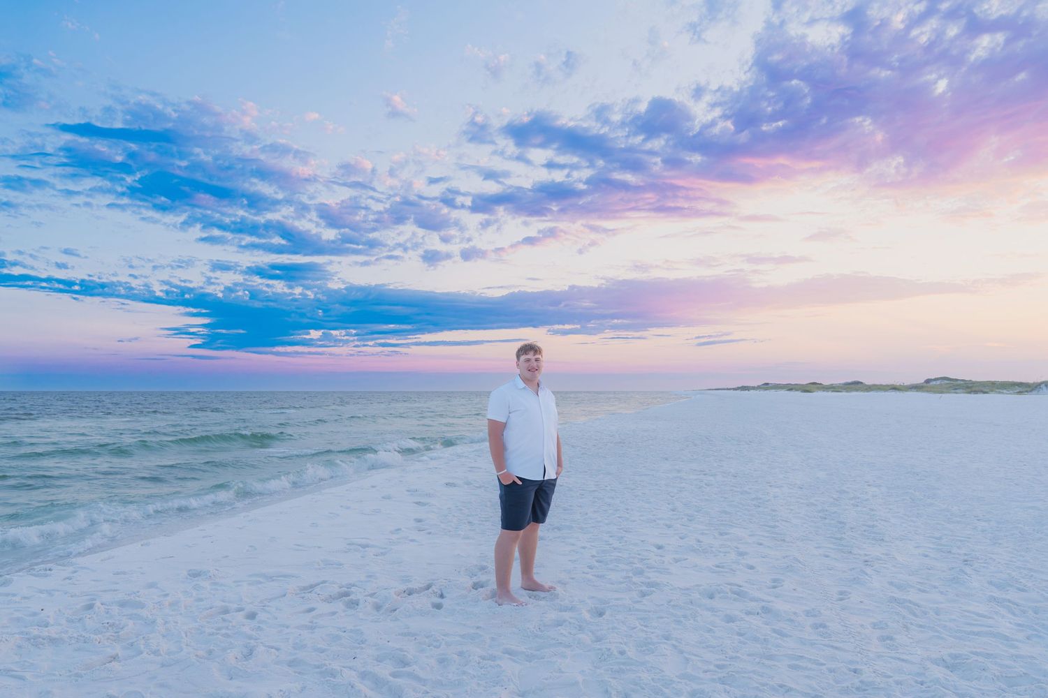 Man standing on white sand beach at sunset with pastel pink and blue sky over calm ocean waters.
