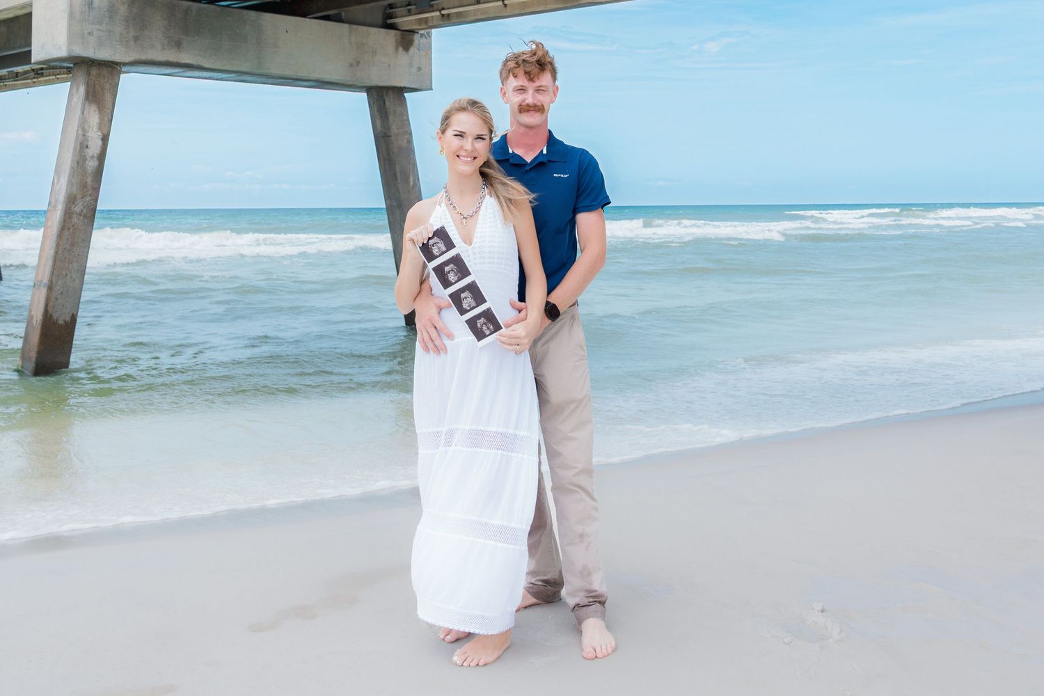 Couple on beach holding ultrasound photos, woman in white dress, man in navy shirt, pier and ocean in background.