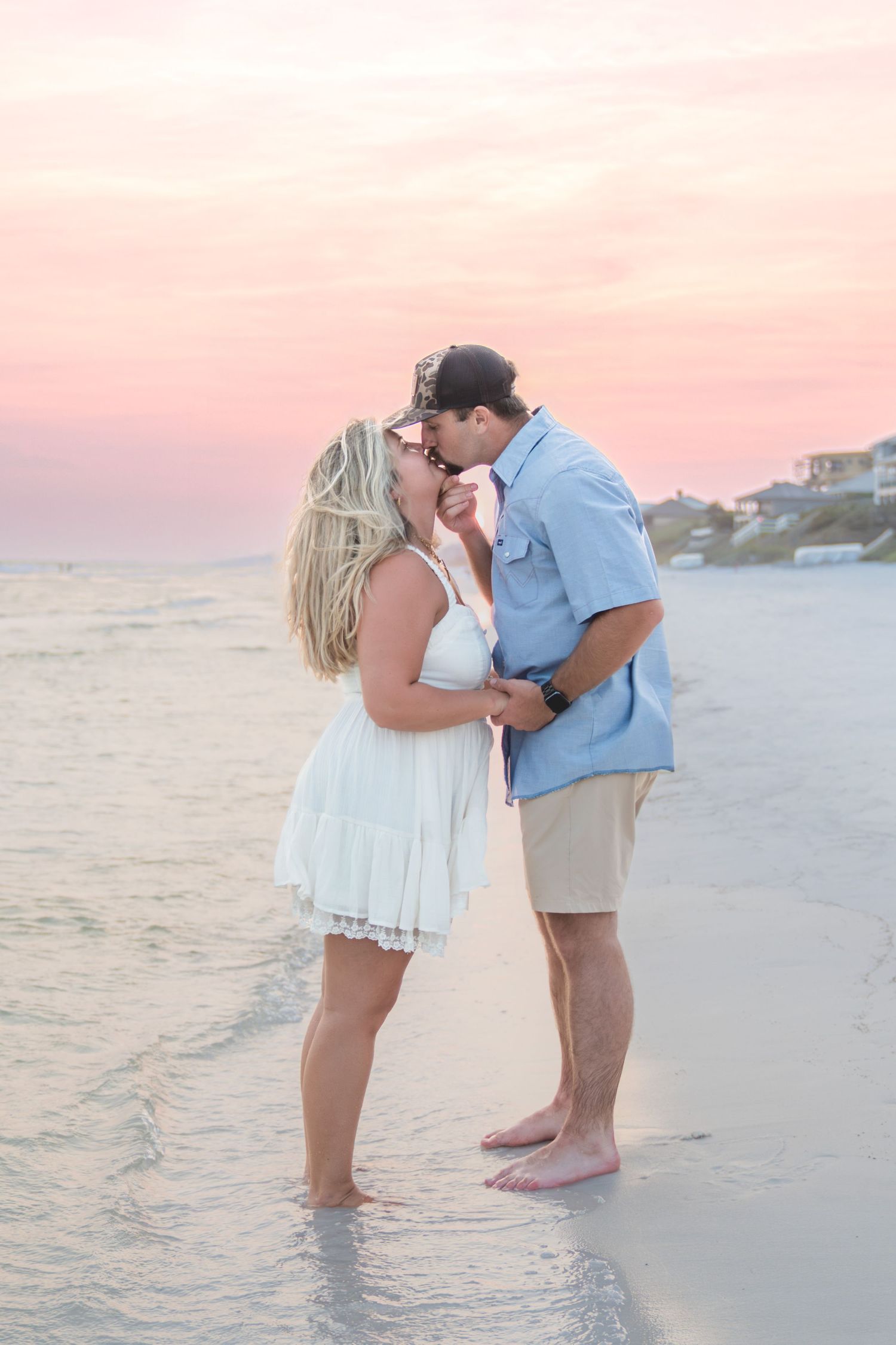 Couple sharing a romantic kiss on a sandy beach at sunset, woman in white dress, man in blue shirt and khaki shorts.