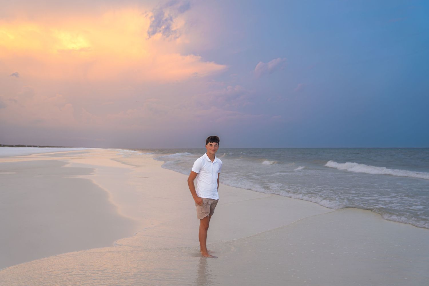 Man in white shirt standing on a serene sandy beach at sunset with colorful sky and ocean waves.