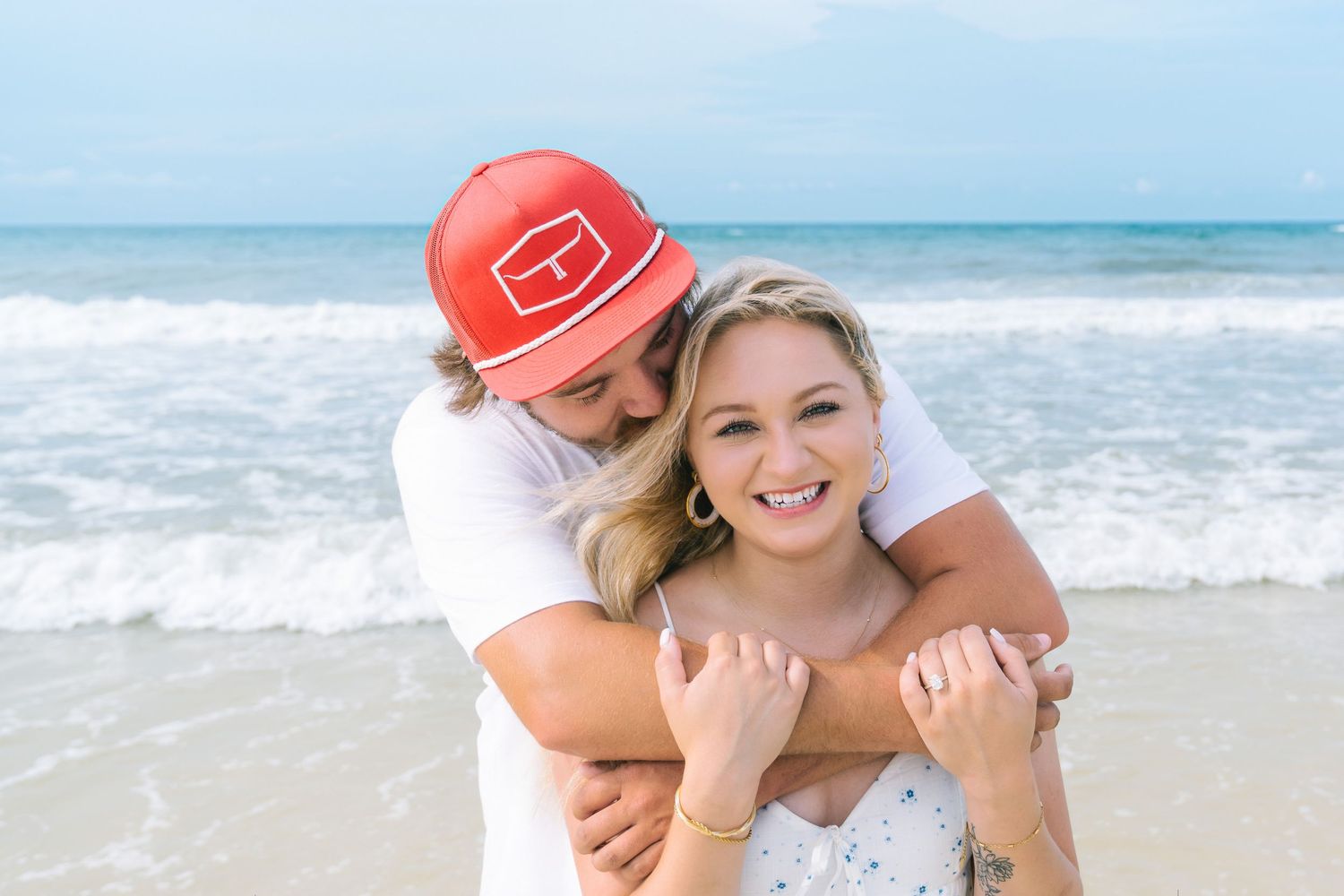 Couple embracing and smiling on a sunny beach, man in red cap hugging woman in white dress by ocean waves.