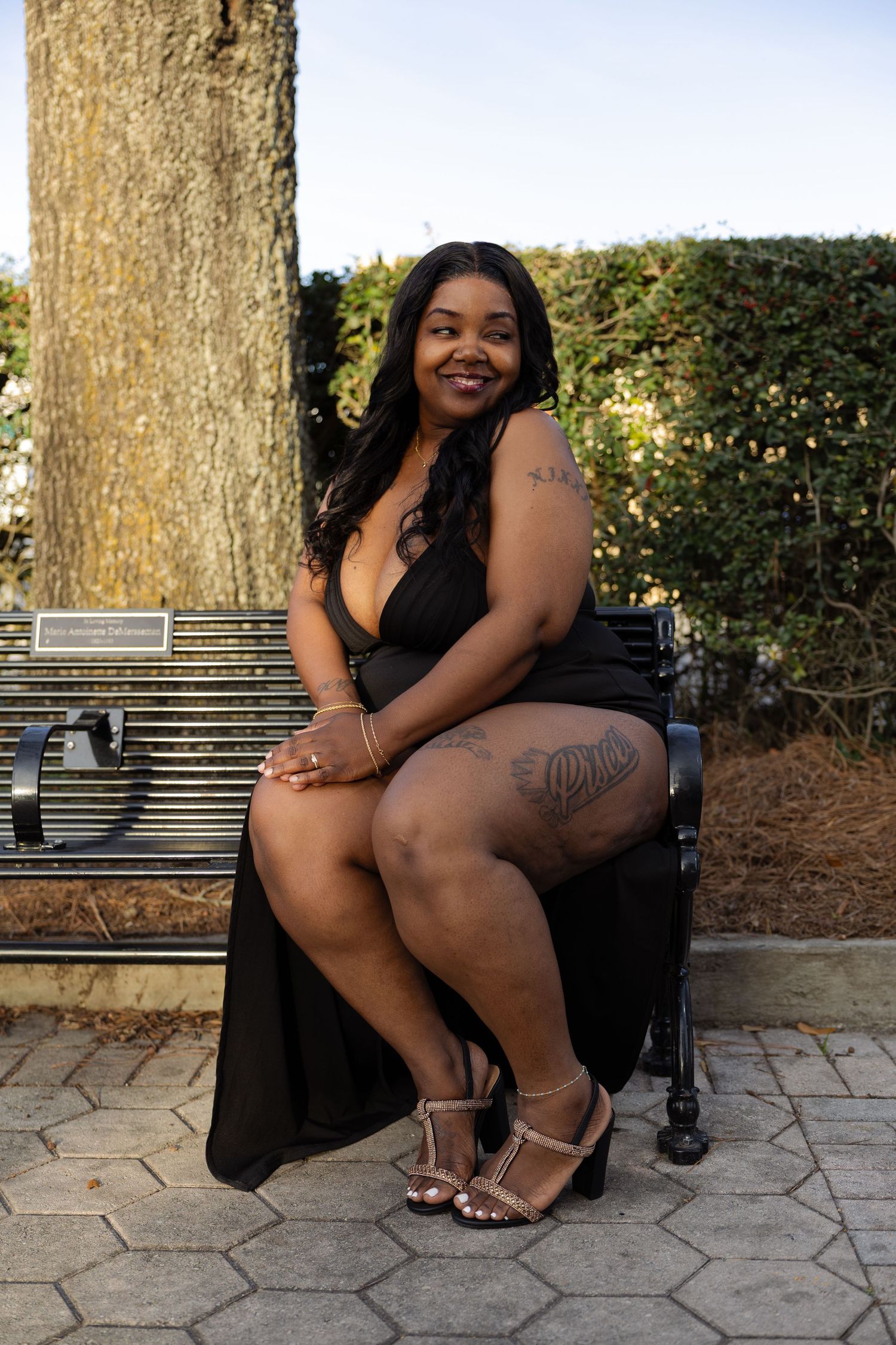 A plus-size woman in a black halter dress and heeled sandals sits on a park bench, smiling, with tattoos visible on her thigh.
