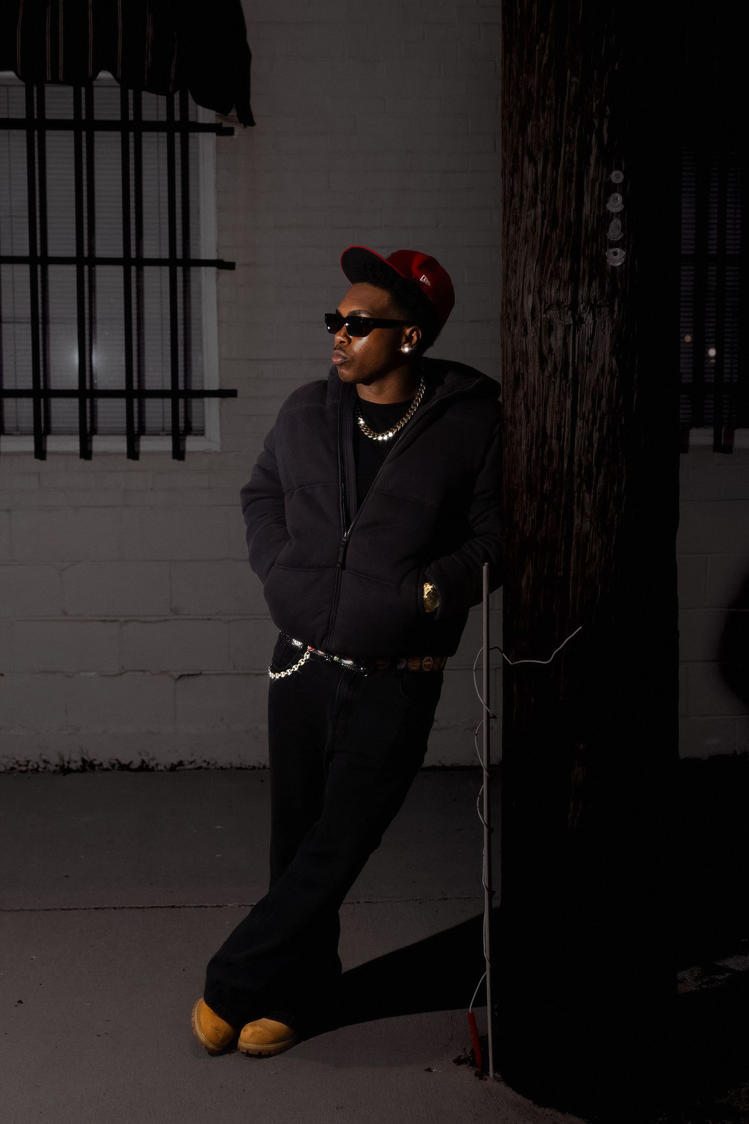 A stylish man in all-black outfit, red cap, and sunglasses leans against a pole outside a building at night.