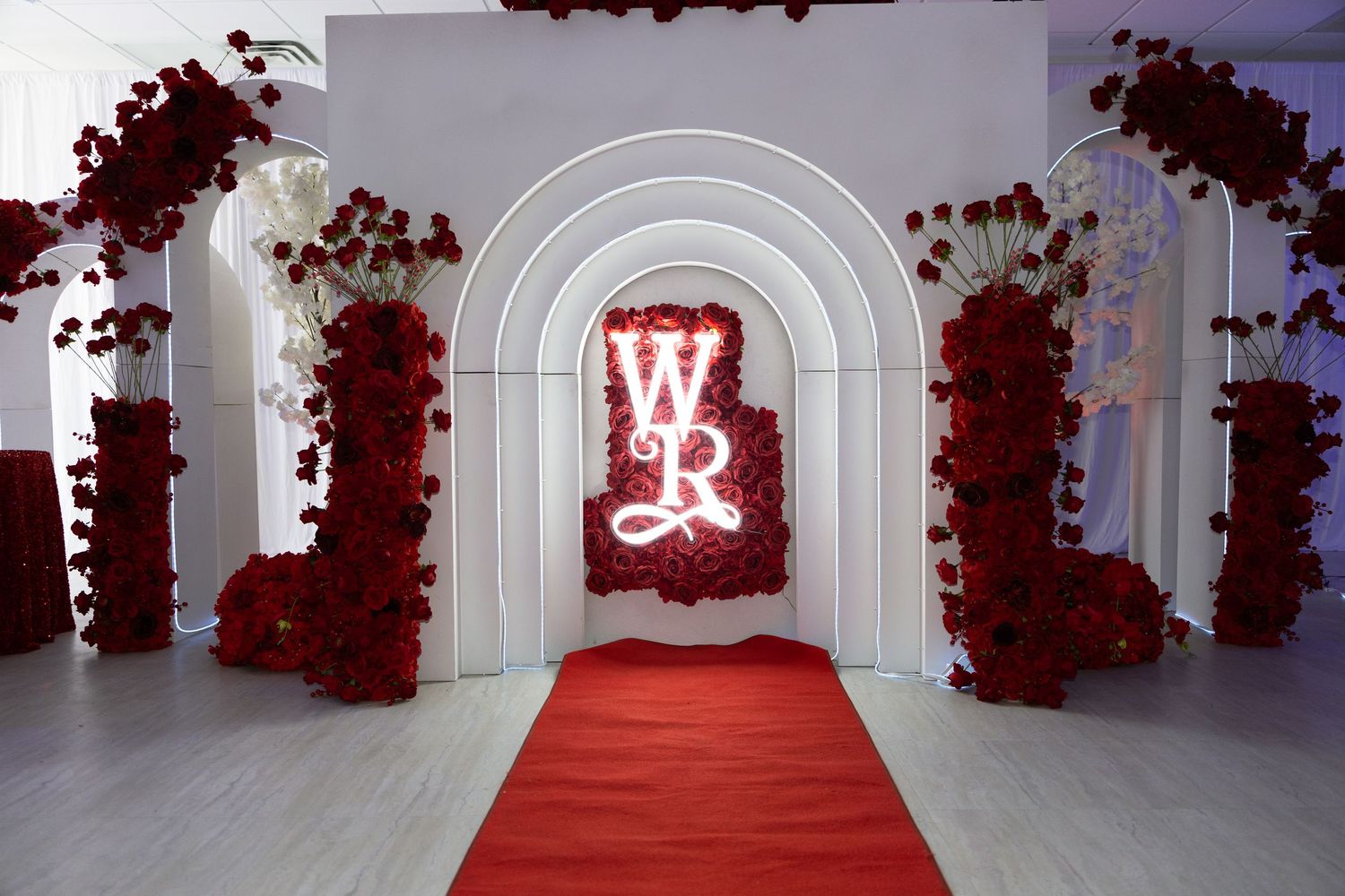 Elegant wedding entrance with red roses, white arches, red carpet, and illuminated WR monogram neon sign.