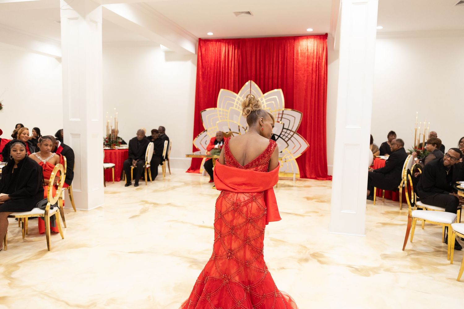 Woman in red lace gown walks runway at elegant fashion show with red draped backdrop and seated audience.