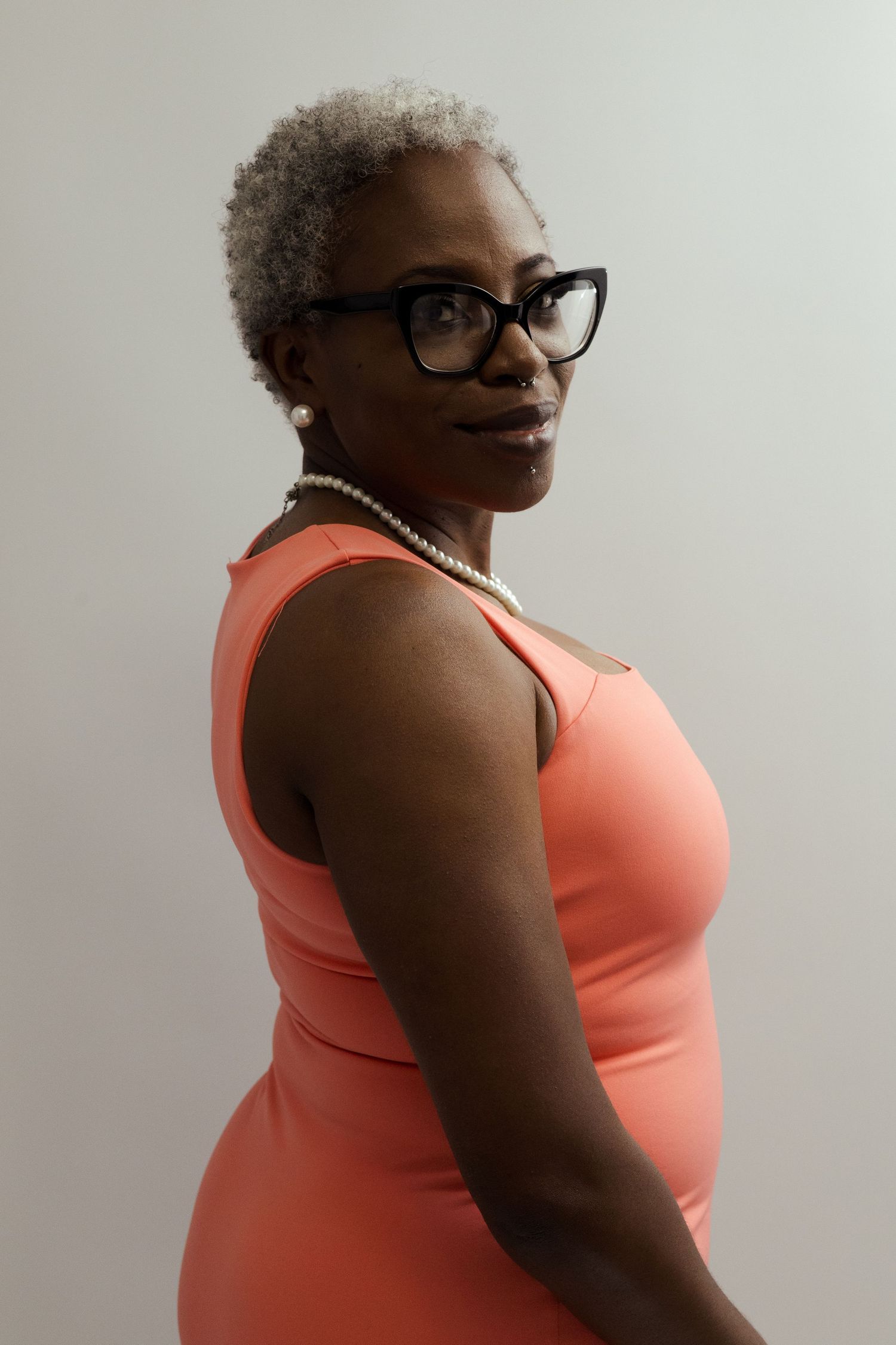 Mature Black woman with gray hair, glasses, pearl necklace, wearing coral dress, posing against light background.