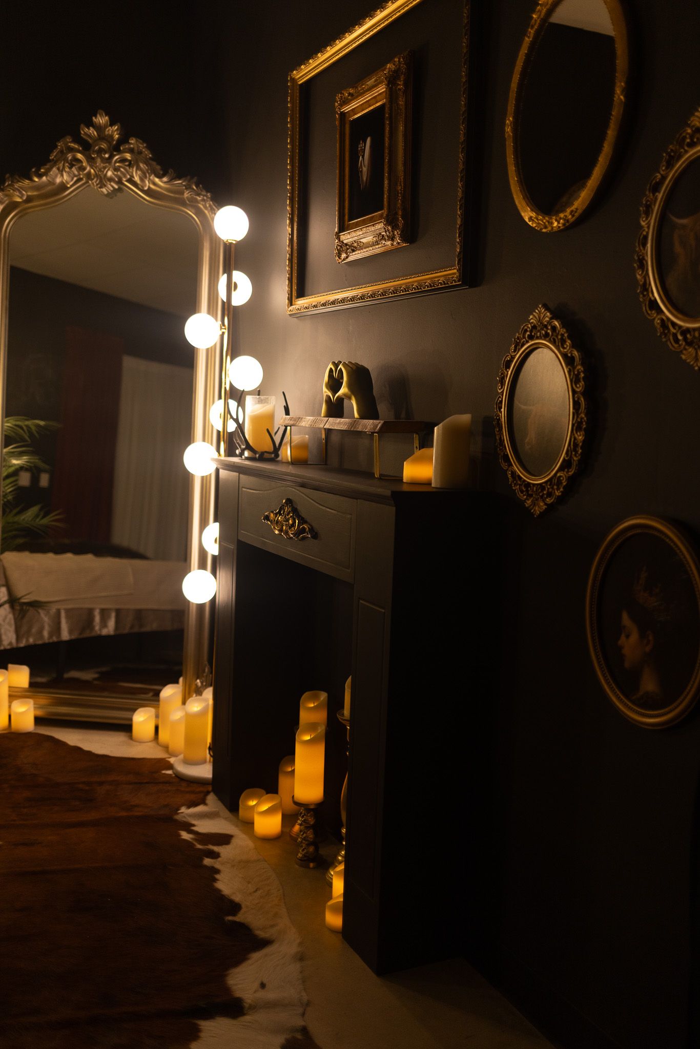 Dark moody room with gold-framed mirrors, lit candles, and a black fireplace with Hollywood vanity mirror.