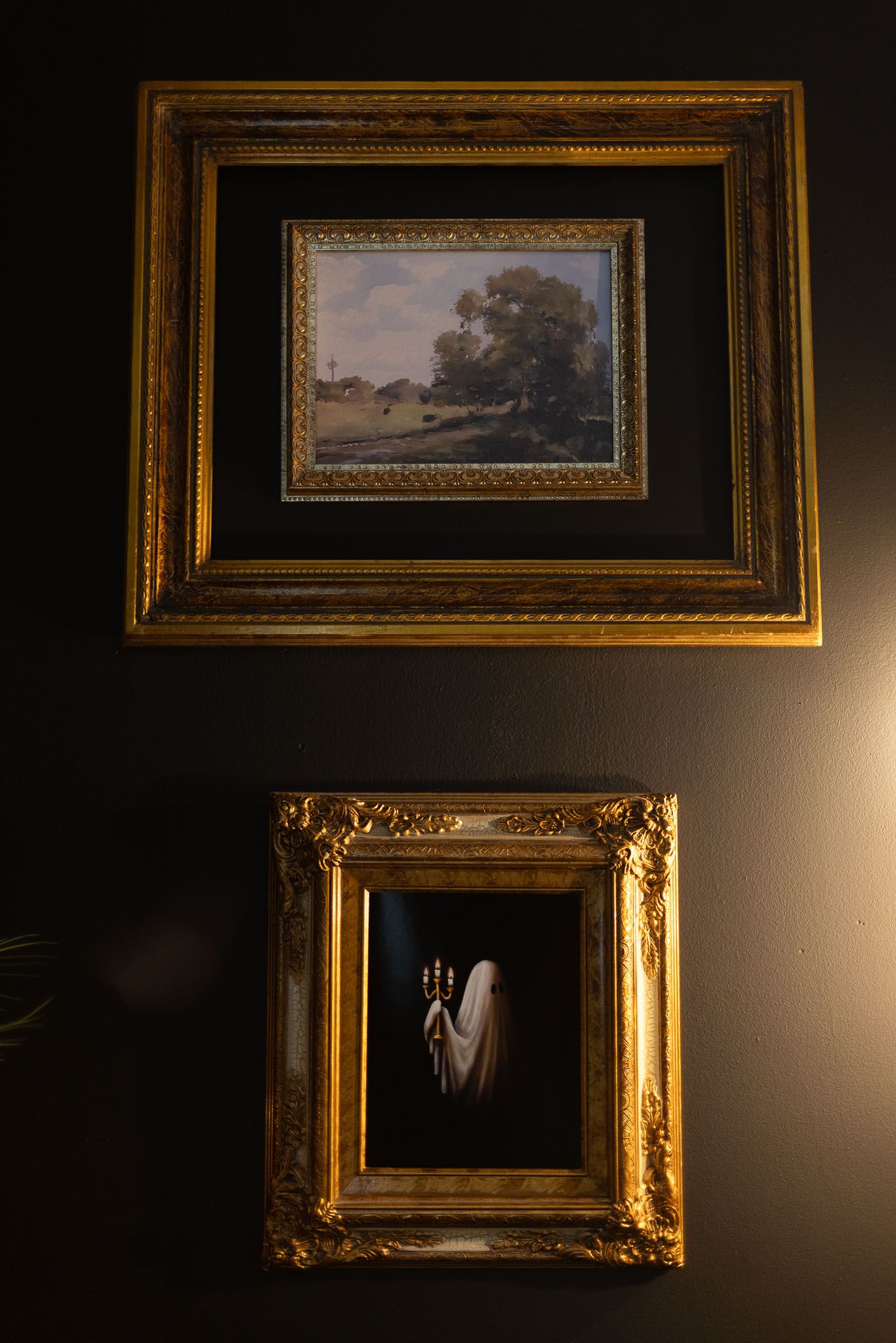 Two ornate gold-framed artworks on a dark wall, featuring a landscape painting above and a moody portrait below.