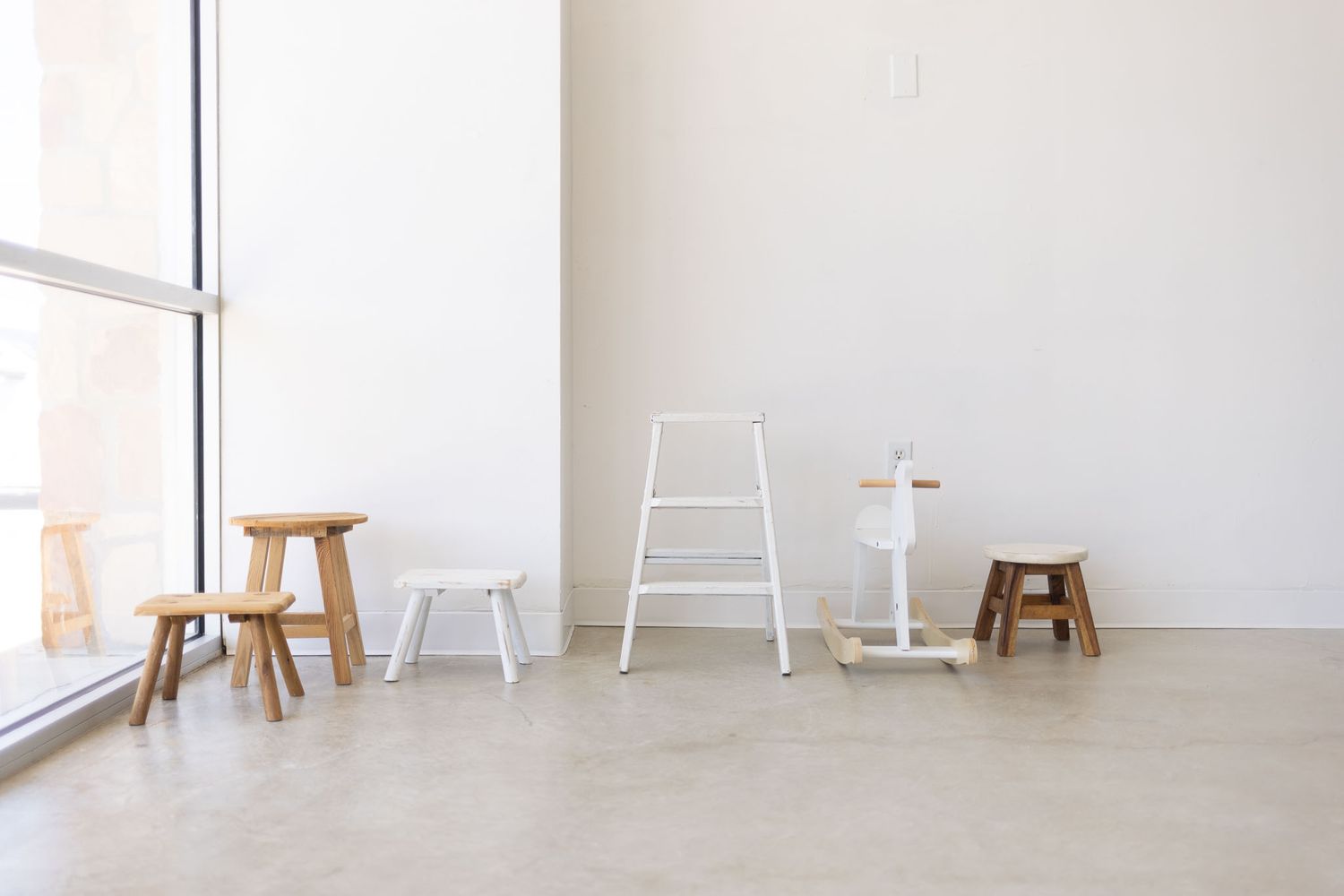 Minimalist white studio space with wooden stools, white ladder and table against blank wall near bright window.