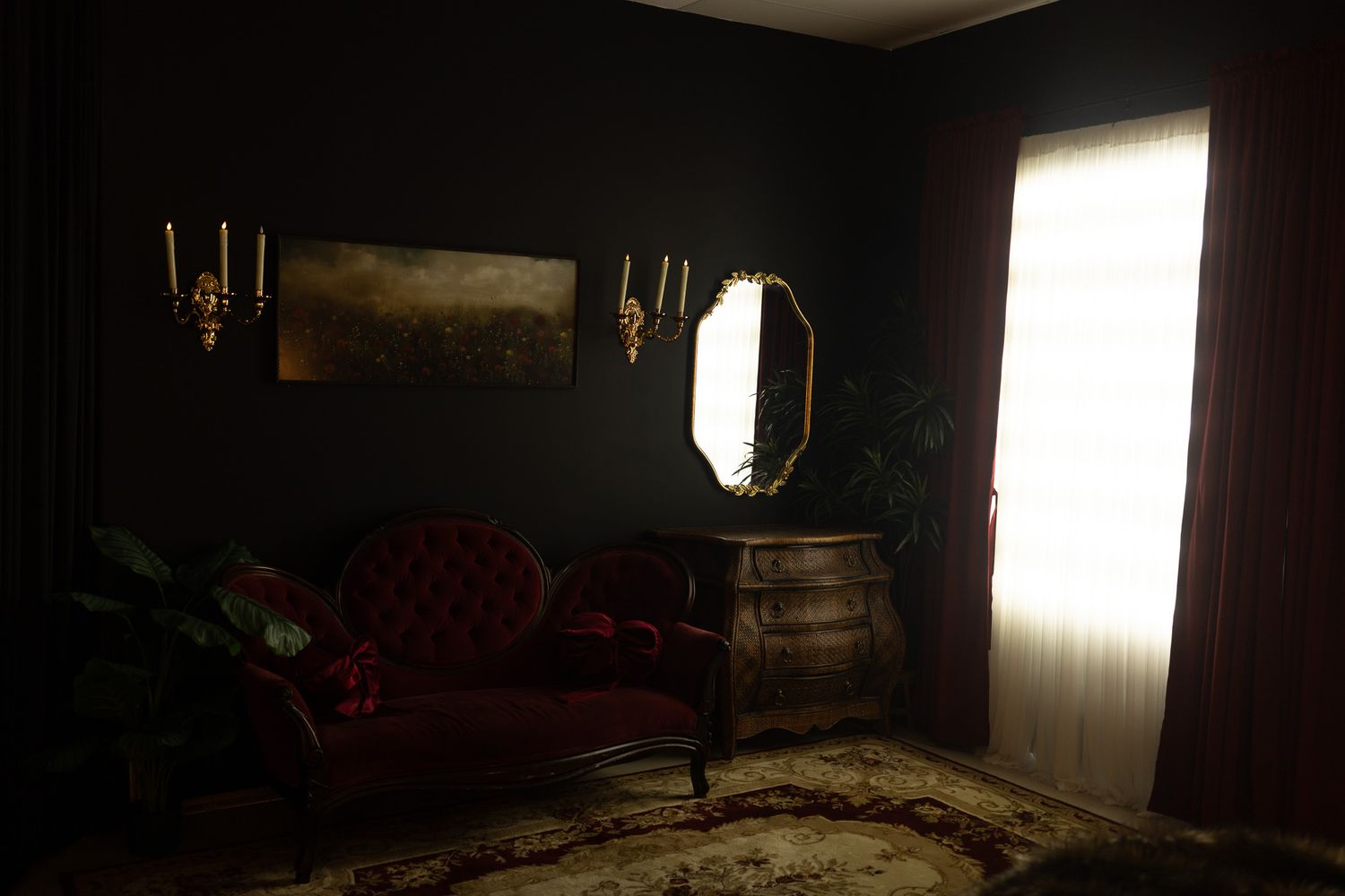 Dark gothic living room with red velvet sofa, ornate mirror, brass candle sconces, and dramatic black walls.