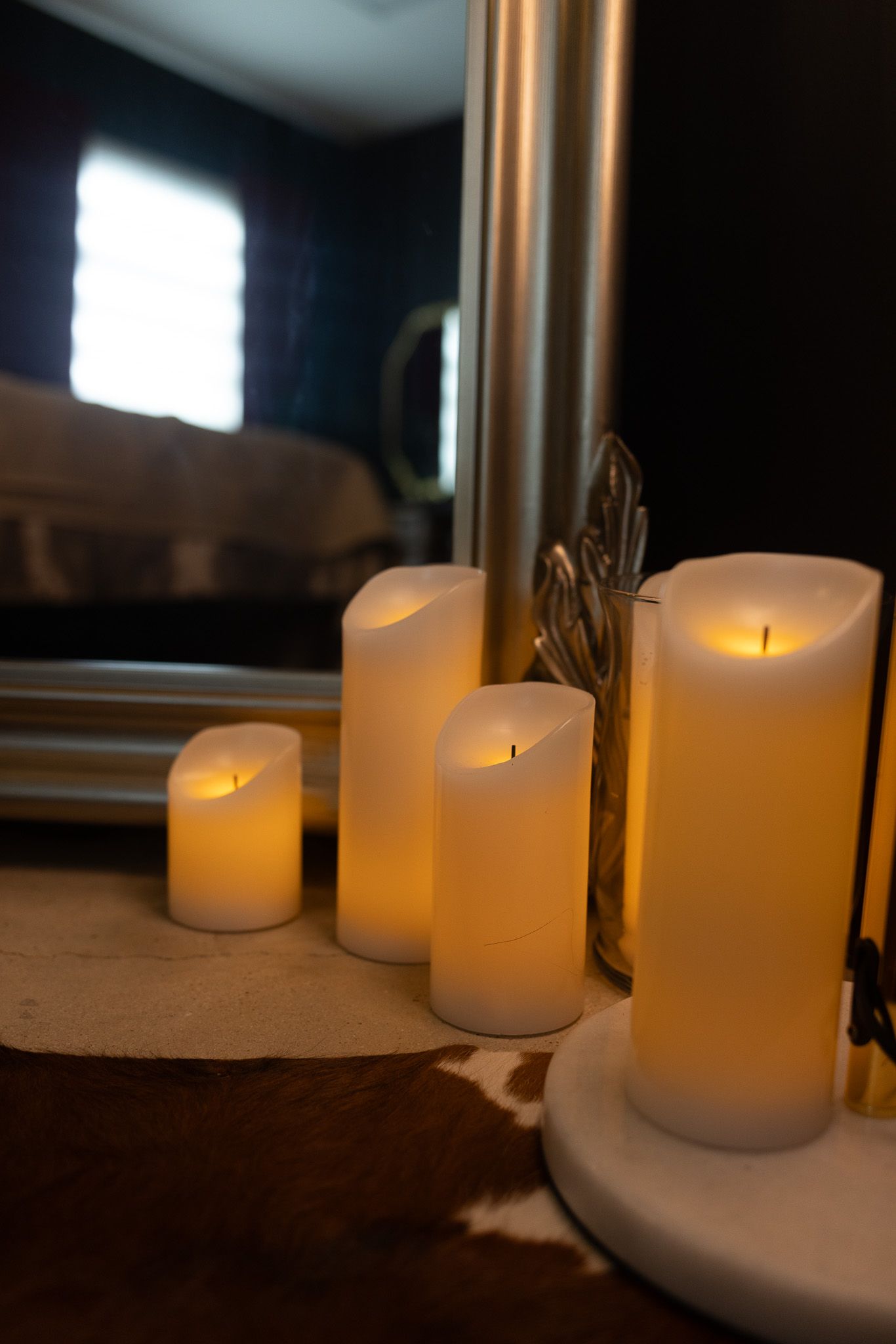 Four glowing white pillar candles arranged by a mirror on a wooden surface in a dimly lit room.