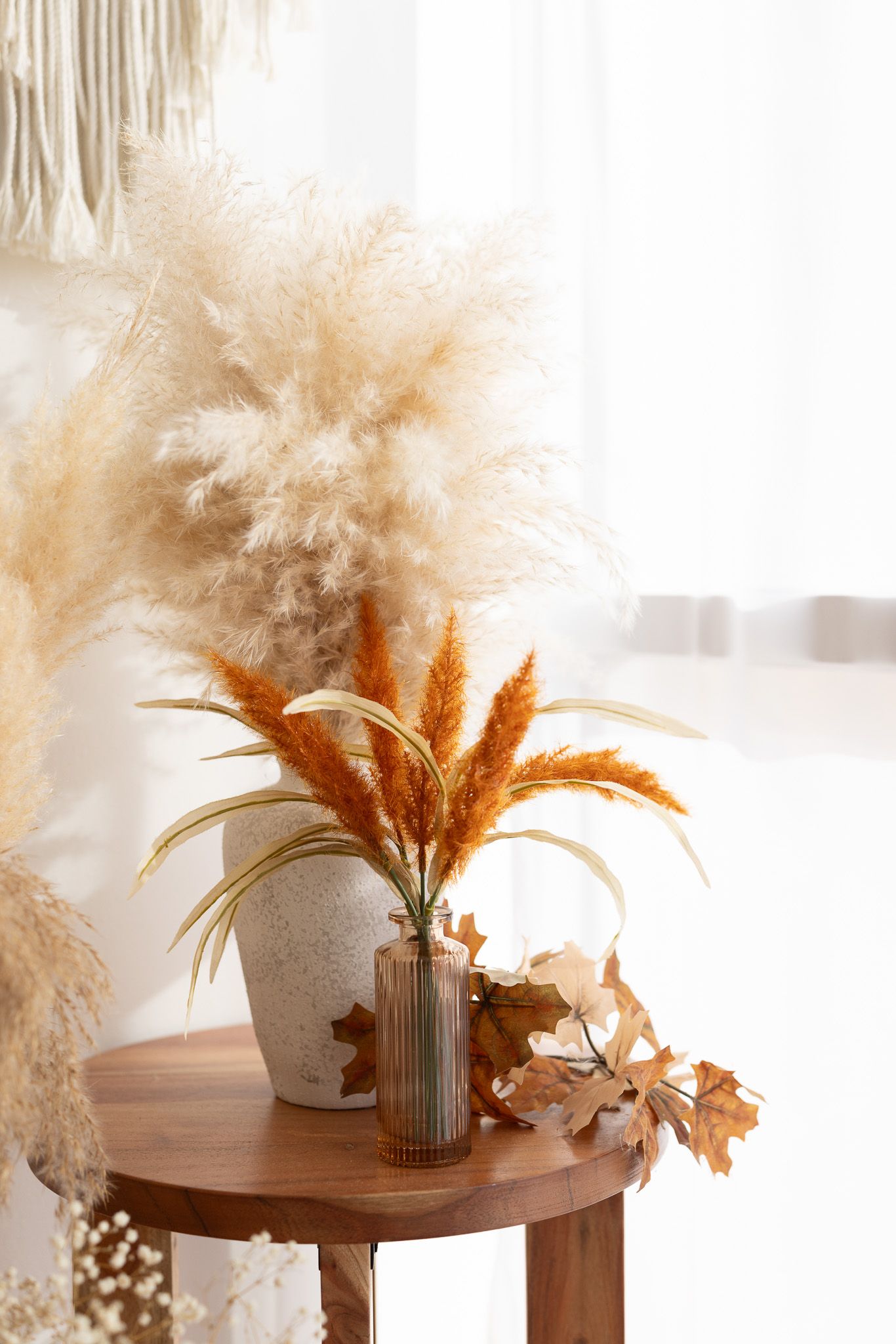 Boho fall decor with pampas grass and dried leaves in stone vase on wooden table near white curtains.