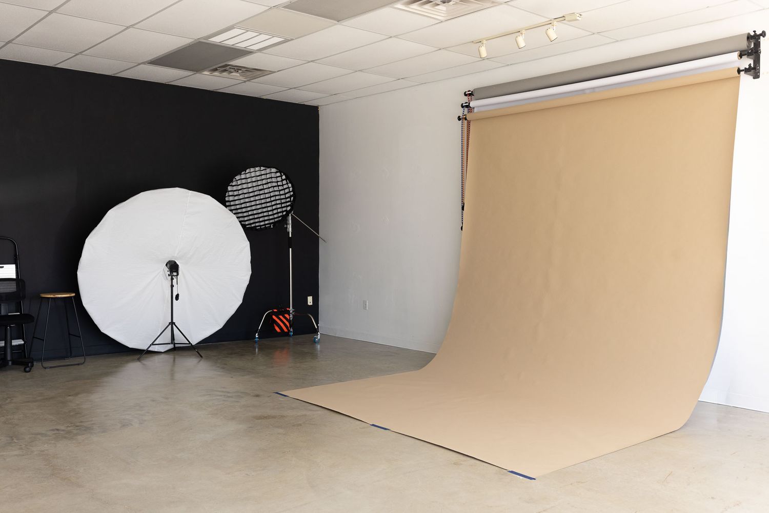 Professional photography studio setup with white reflector, light stand, and seamless tan backdrop against black and white walls.