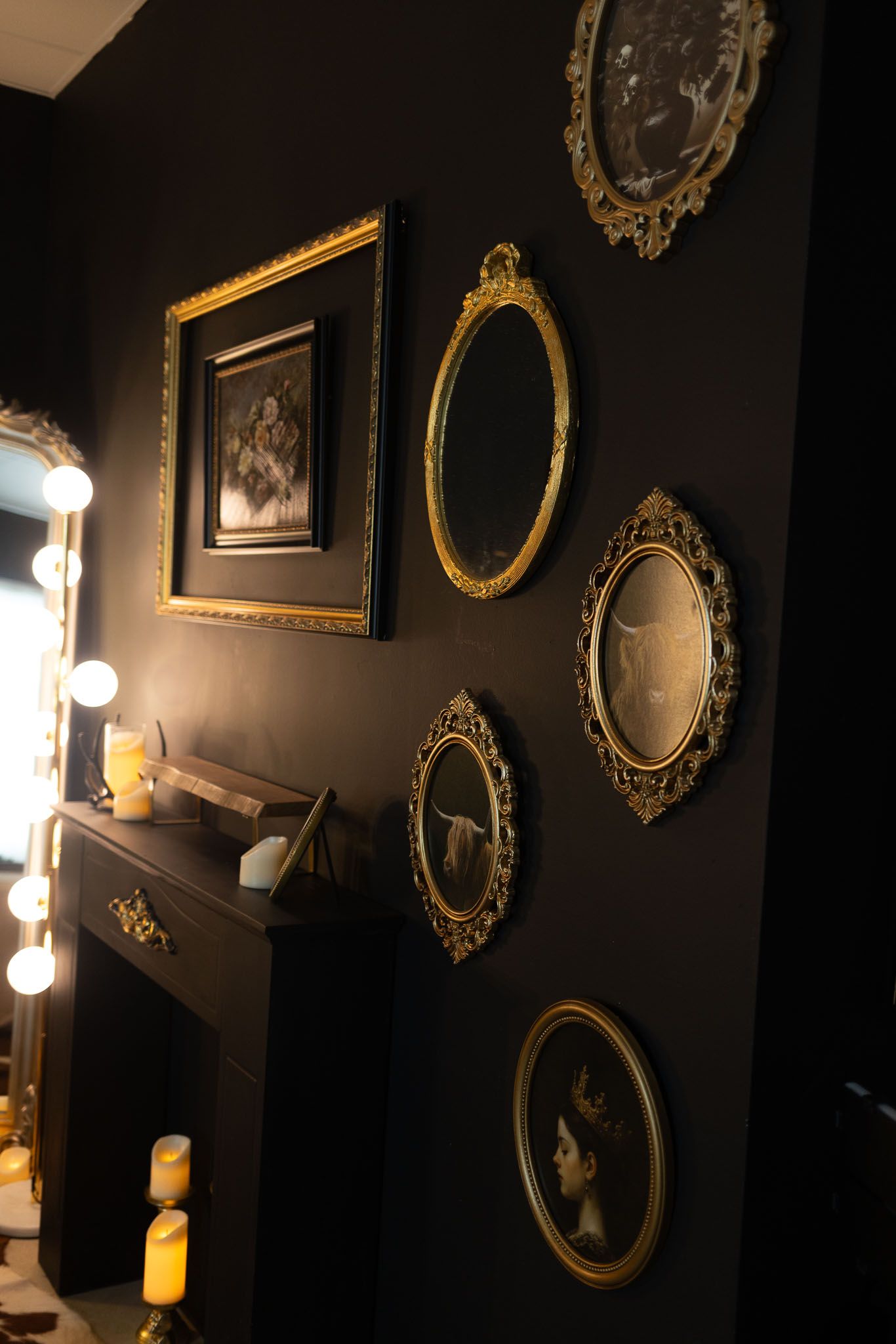 Vintage gold-framed mirrors and artwork arranged on a dark wall beside a vanity with illuminated makeup lights.