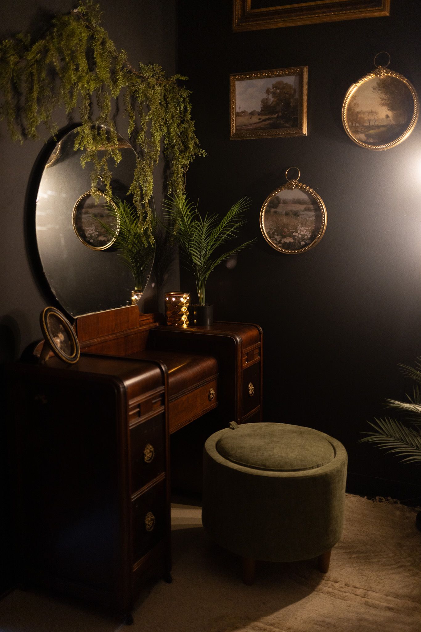 Dark vintage vanity with round mirror, green velvet ottoman, framed wall art, and lush plants in moody dark room.