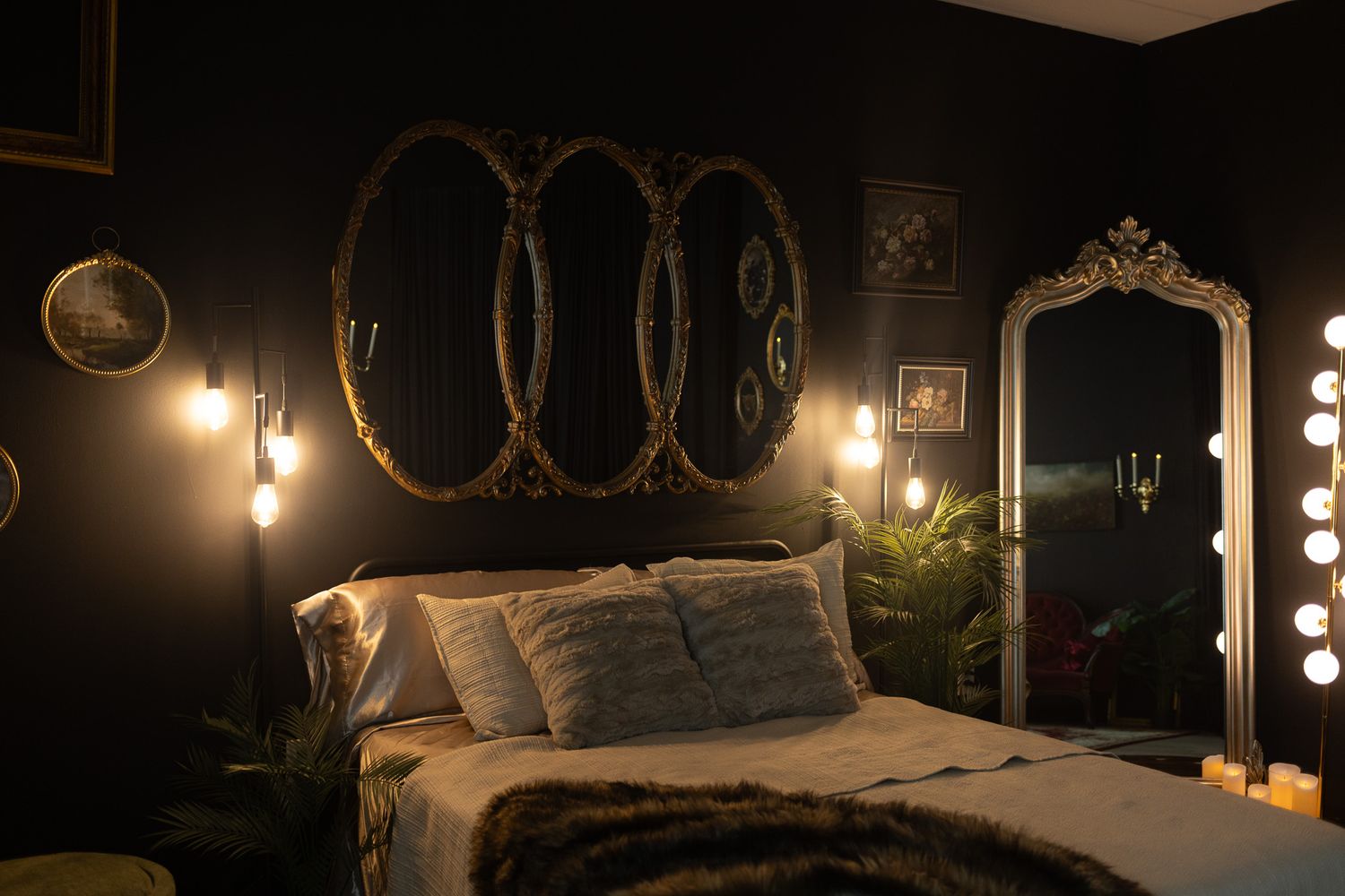 Moody dark bedroom with gold oval mirrors, warm Edison bulb lighting, fur throw, and an ornate full-length mirror.
