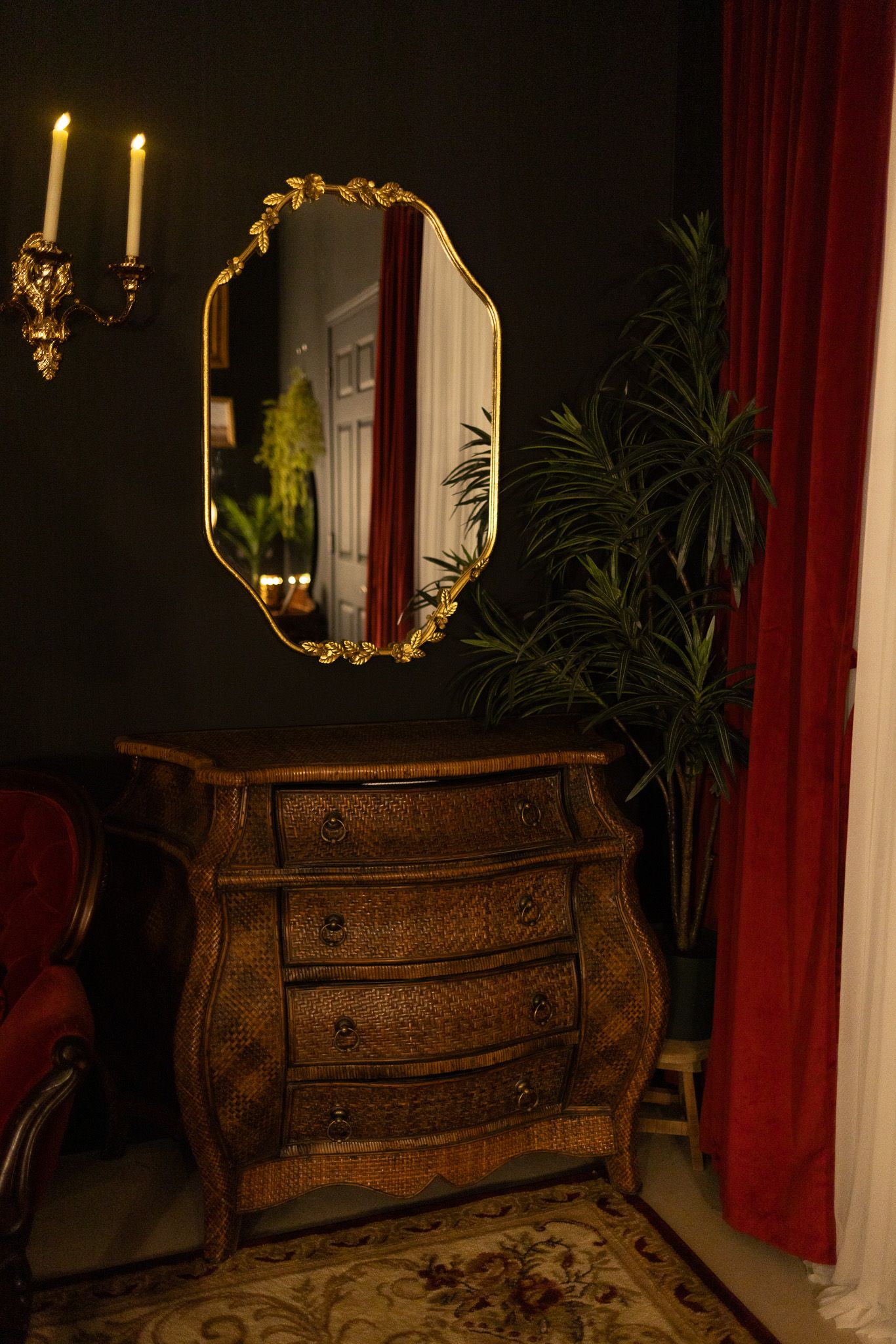 Ornate gold-framed mirror above antique wooden dresser with dark walls, red curtains, candle sconce, and tropical plant.