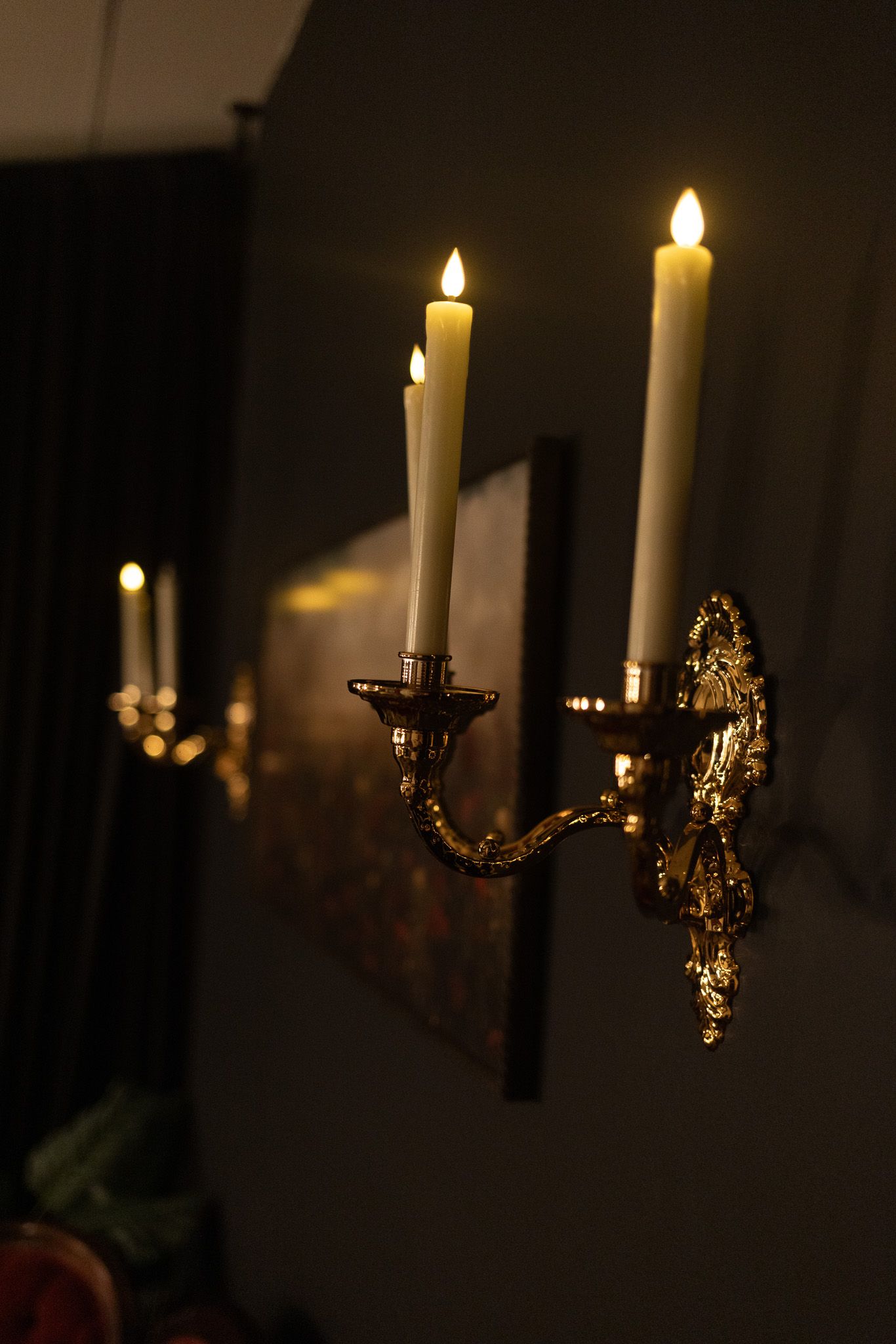 Gold brass wall sconce with three lit white taper candles casting warm light in a dark elegant room.