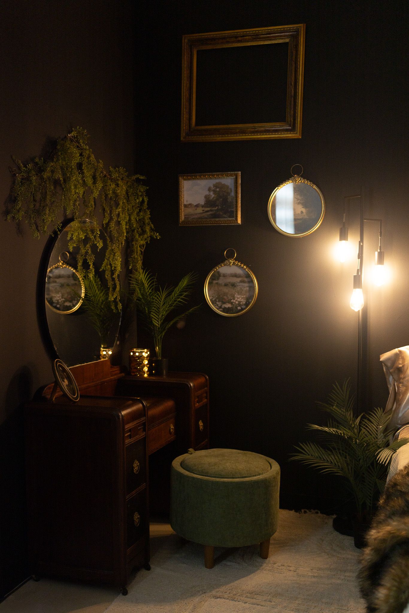 Dark moody bedroom corner with vintage dresser, green velvet ottoman, tropical plants, and gold framed mirrors.