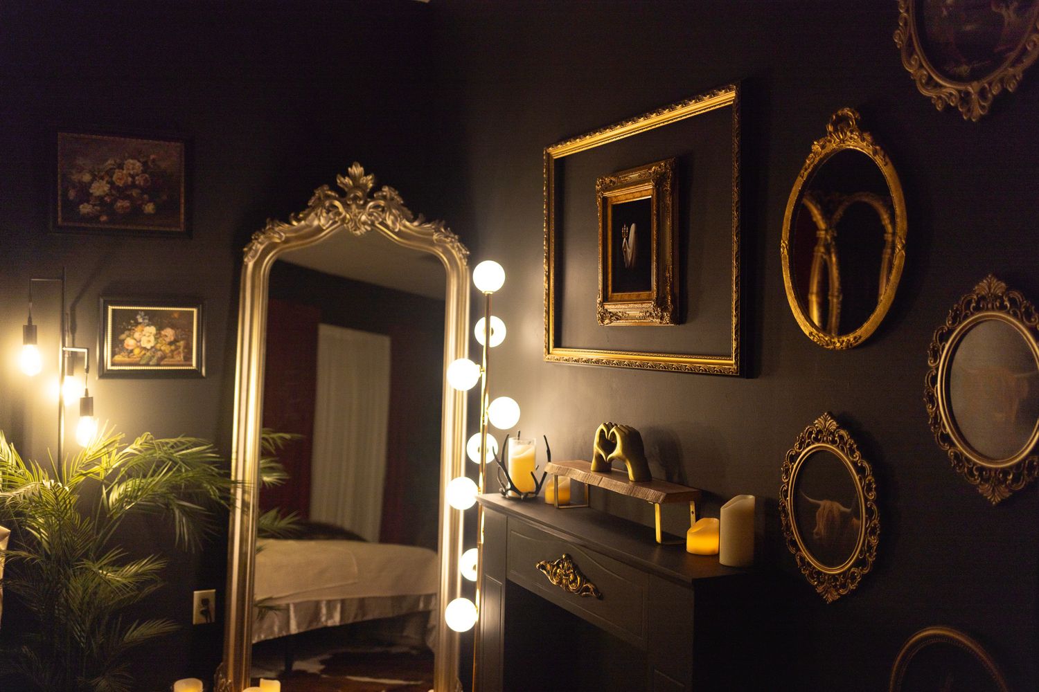 Dark moody bedroom with gold ornate mirrors, Hollywood vanity lights, and black walls creating a glamorous gothic aesthetic.