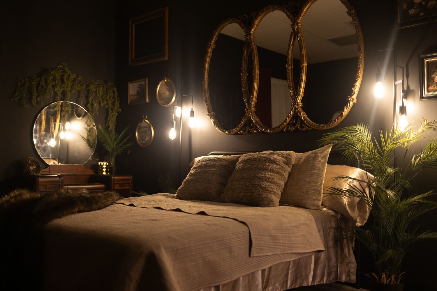 Moody dark bedroom with oval gold mirrors, warm Edison bulb lighting, lush greenery, and neutral bedding for a cozy aesthetic.