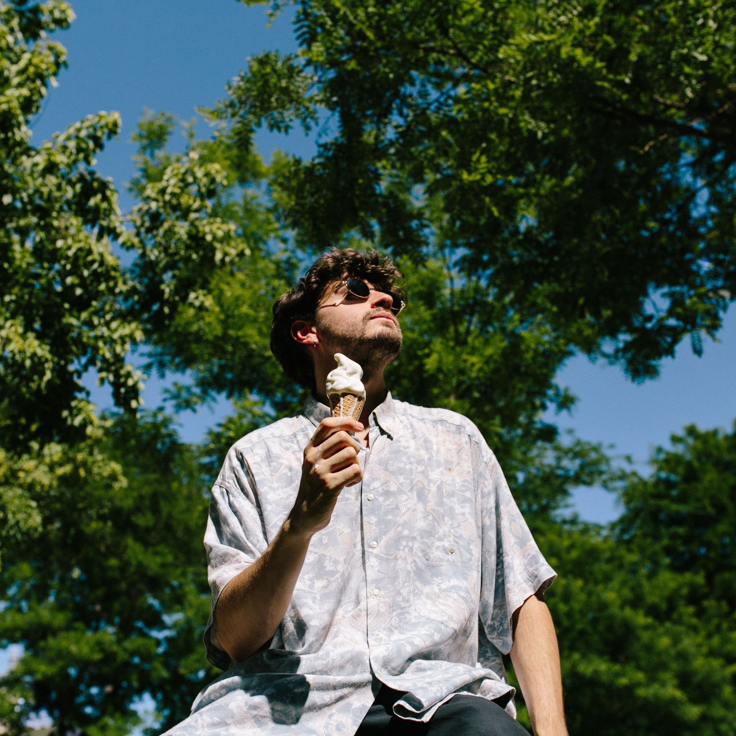Young man holding an ice cream cone looking up while standing under lush green trees in summer.