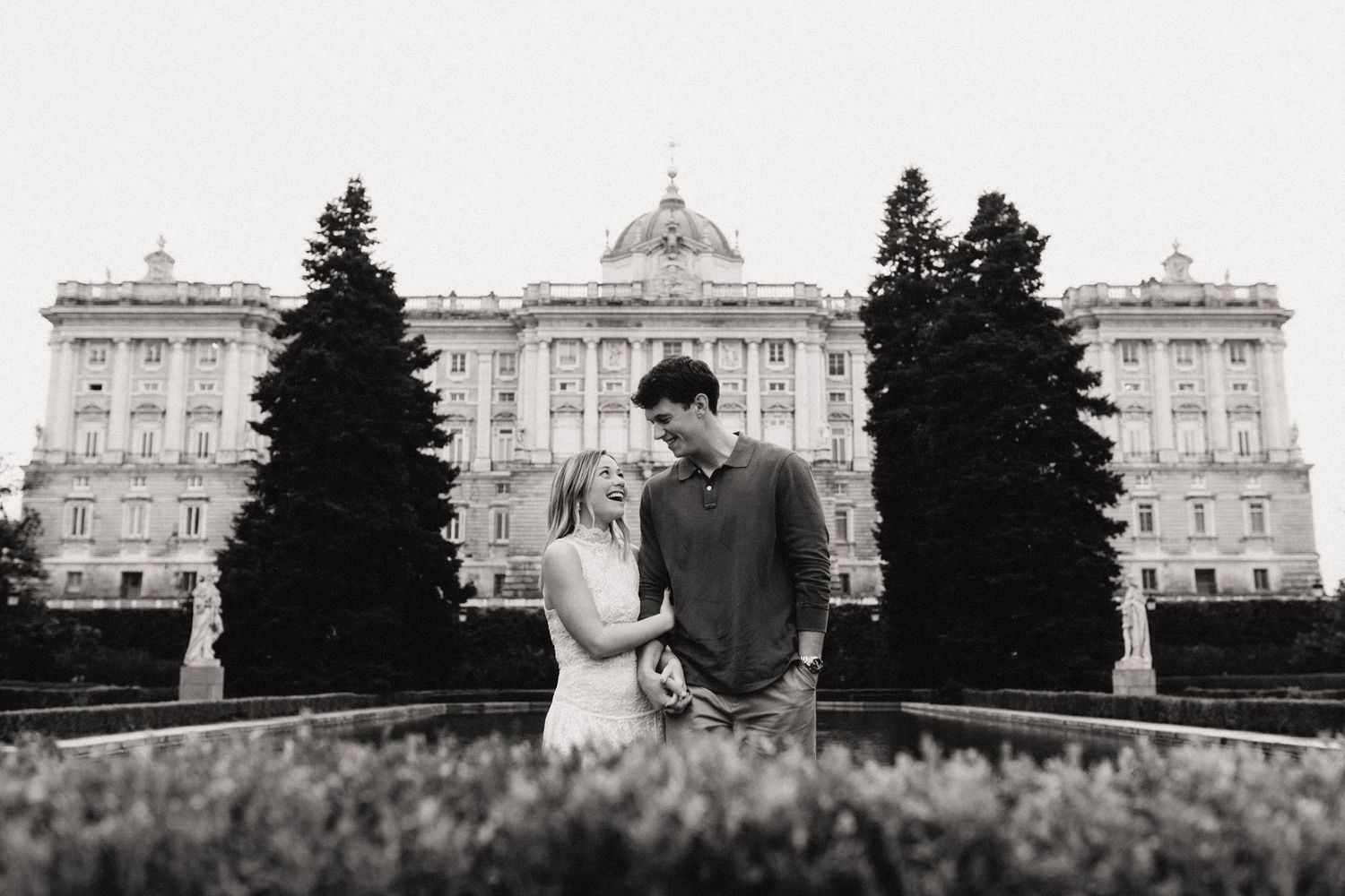 Black and white photo of a couple holding hands before a grand royal palace facade.