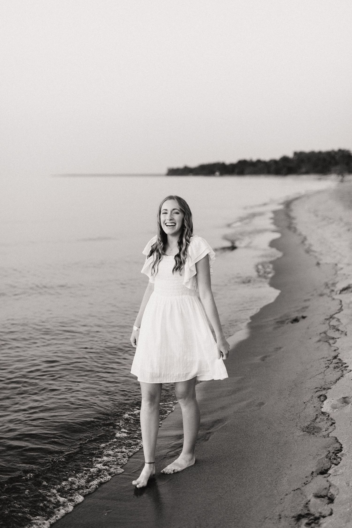 Black and white photo of smiling teenage girl in white dress walking along beach shoreline.