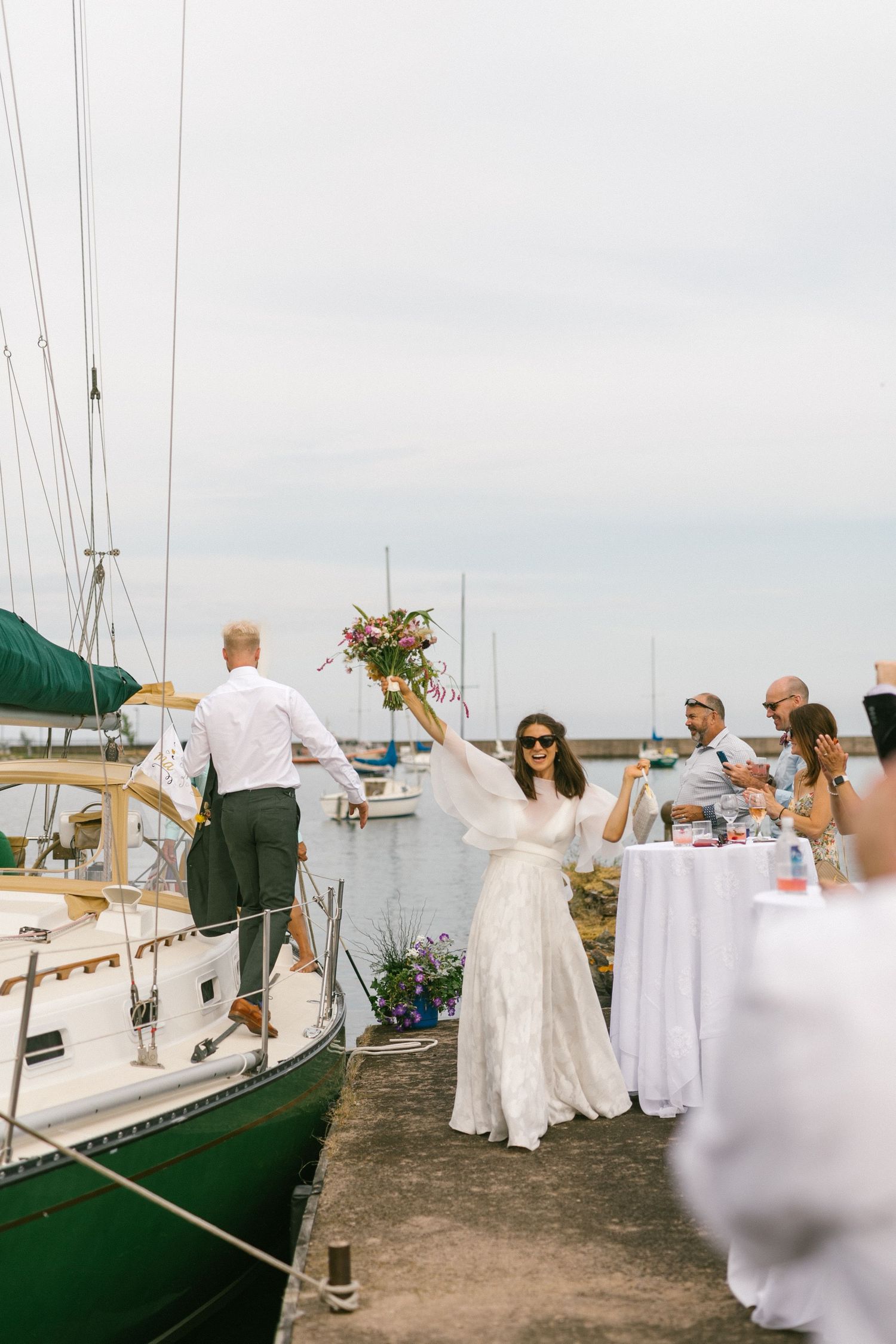 Wedding reception setup with white tablecloths and decor near docked sailboats at a marina.