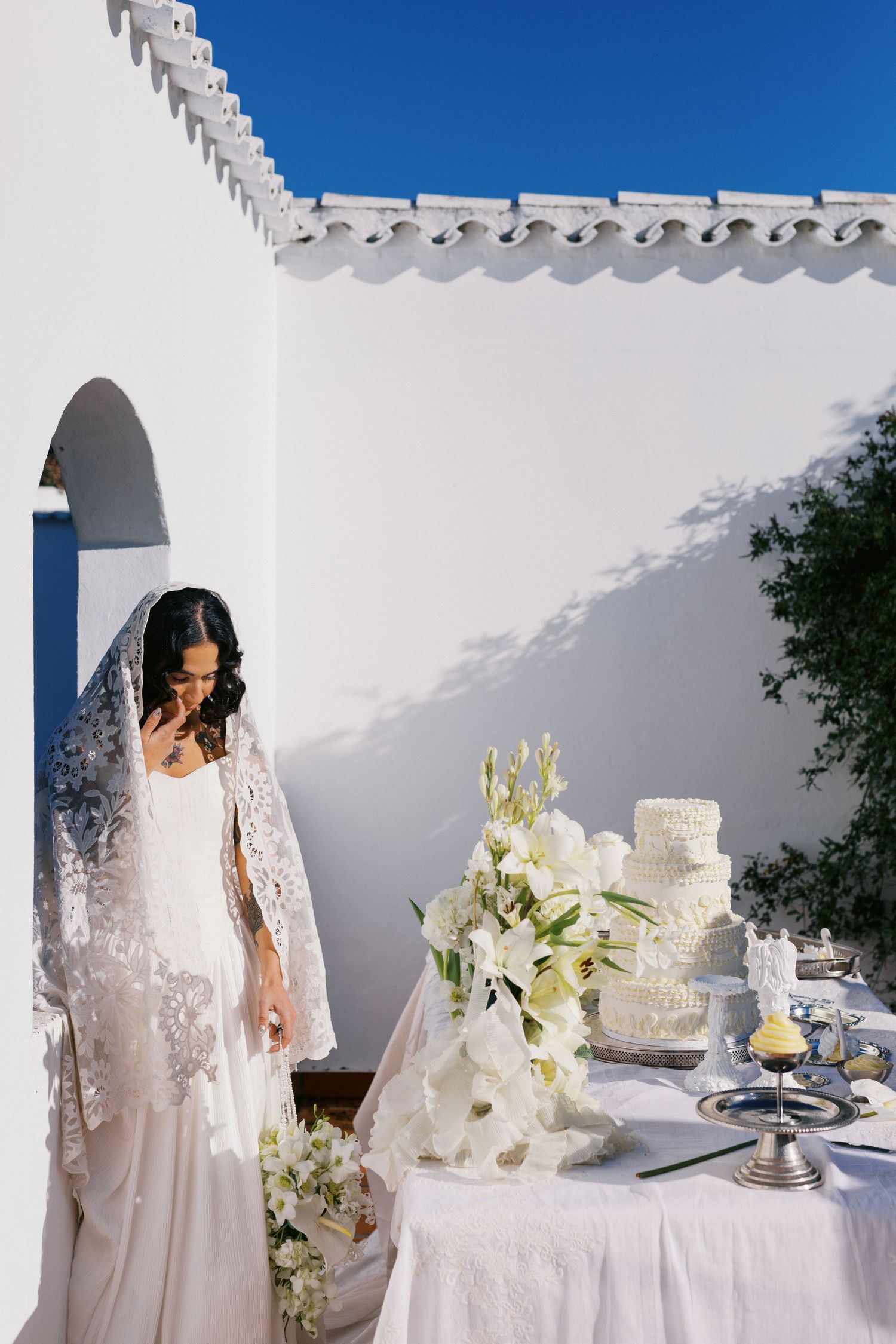 Bride in white lace veil gown stands by elegant wedding cake with white florals against sunlit white Mediterranean wall.