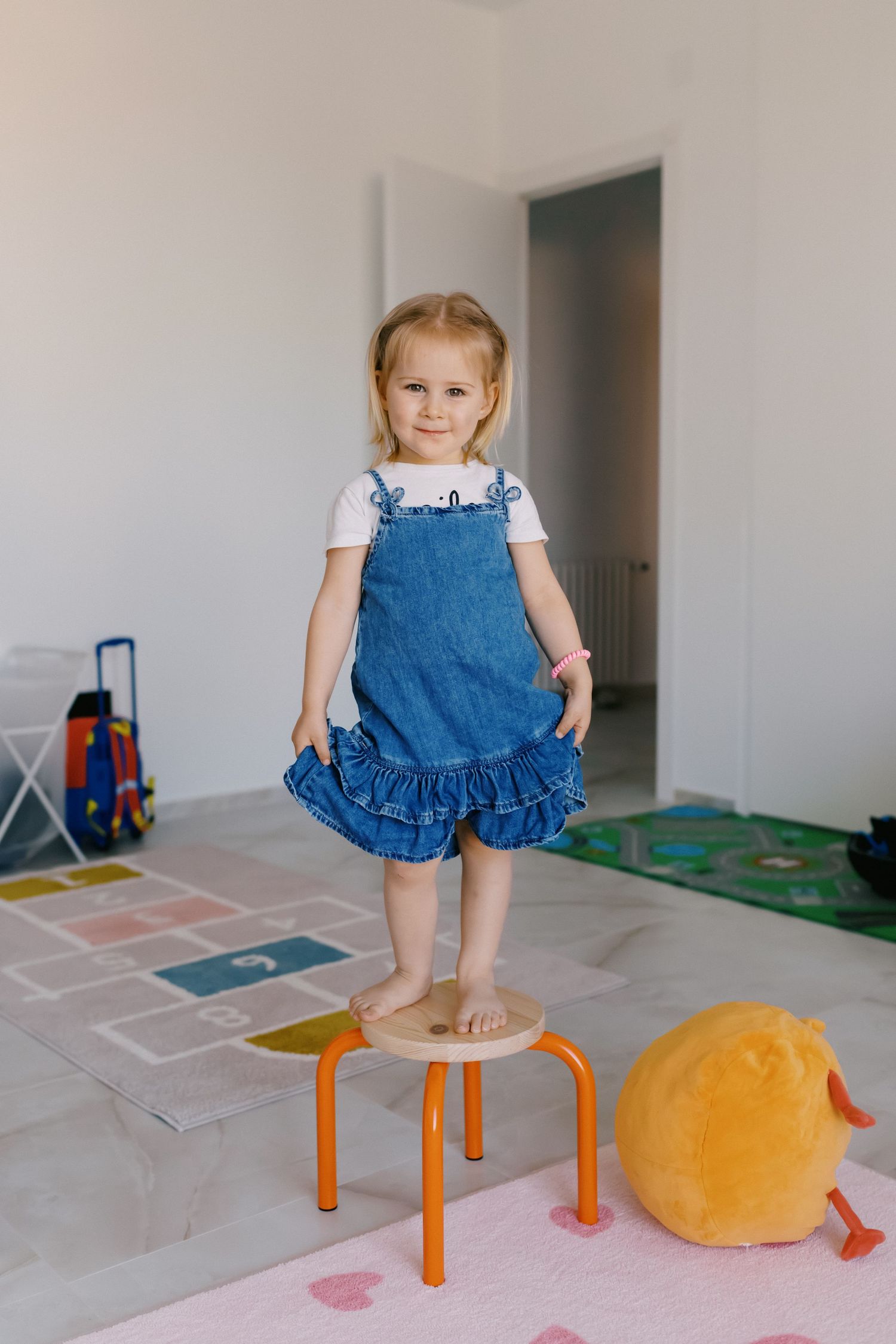 Toddler girl in denim overalls standing on small orange stool in bright playroom with colorful mat.