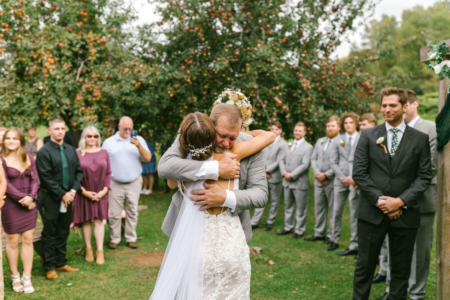 A father embraces his daughter the bride her wedding ceremony in an apple orchard surrounded by guests.