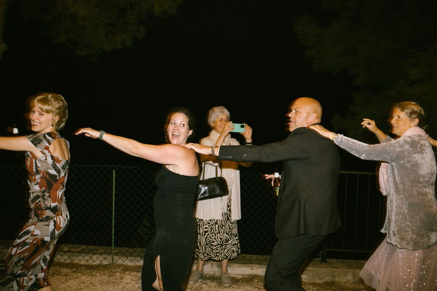 A group of guests dancing in a line together during a nighttime outdoor celebration. Europe and Spain destination wedding photographer.