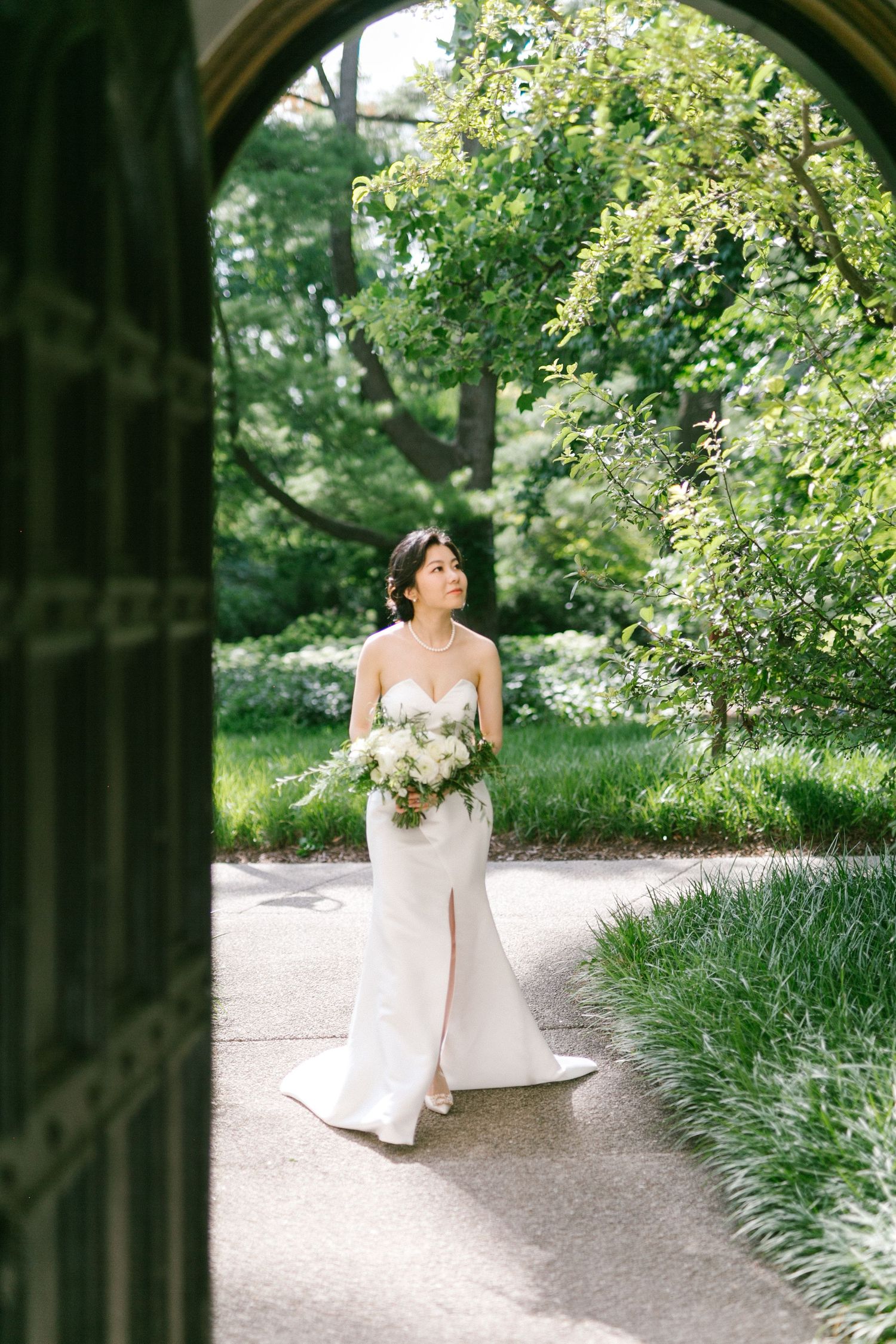 Elegant bride in a strapless white gown holding a bouquet framed by a stone archway in a garden.