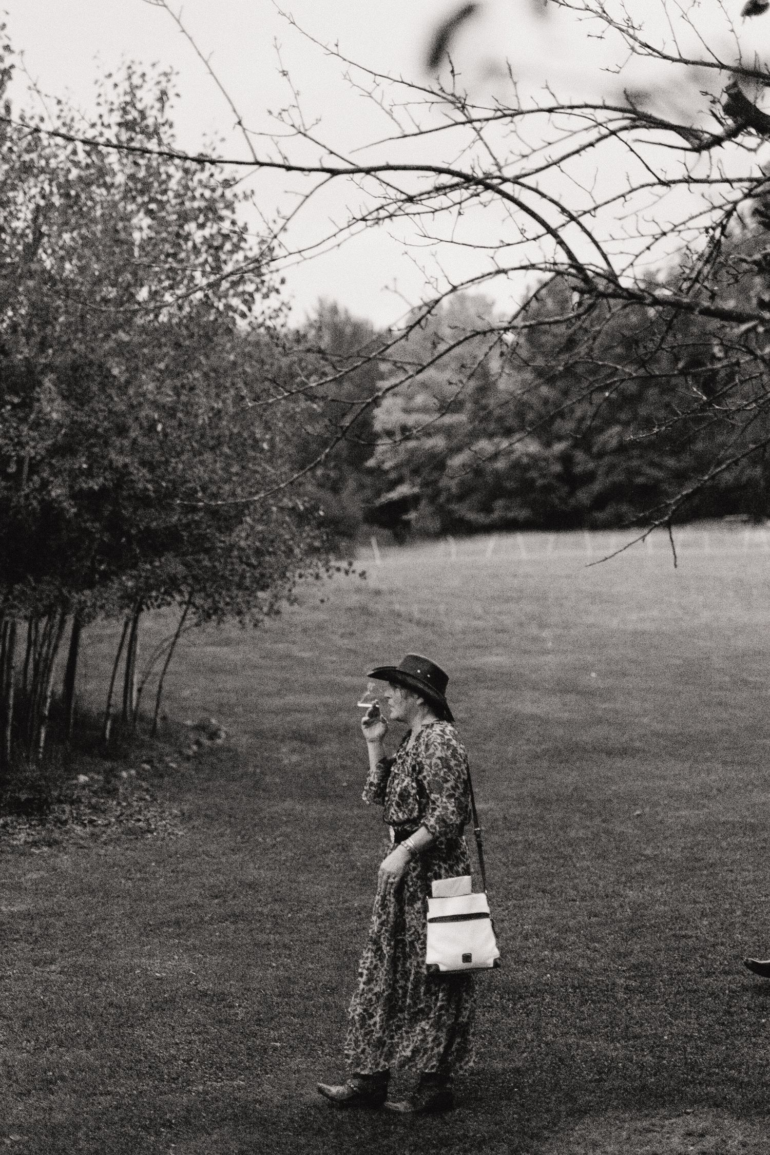 Black and white portrait of stylishly dressed person walking through a field with trees.