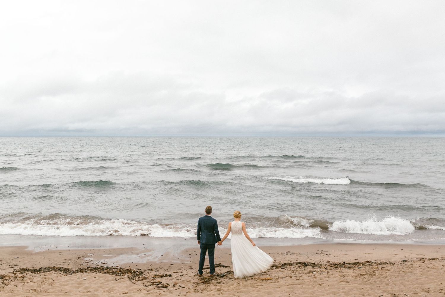 A couple watches the crashing waves on a moody beach shore beneath cloudy skies.