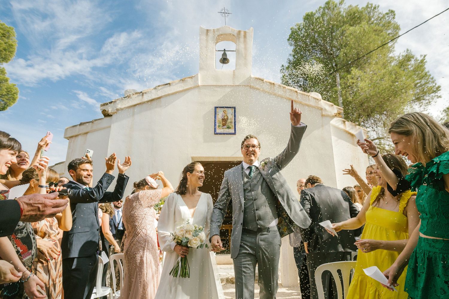 A joyous celebration takes place outside a white Mediterranean chapel with a bell tower on a sunny day. Europe and Spain destination wedding