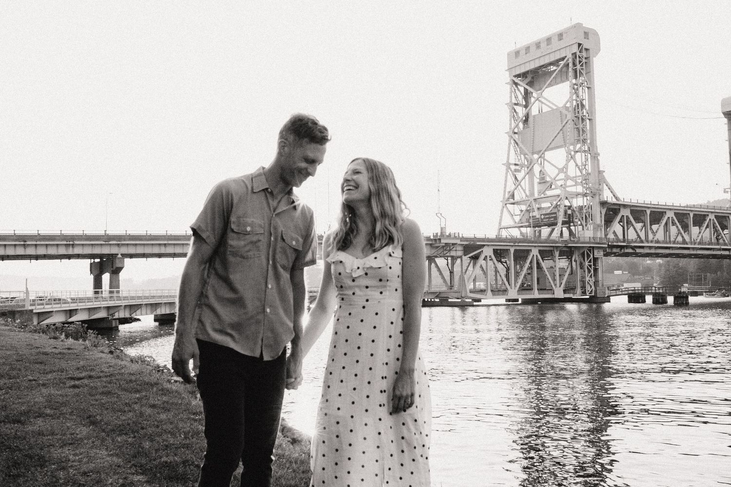 A couple stands near a large bridge on the waterfront as they enjoy a romantic moment together.
