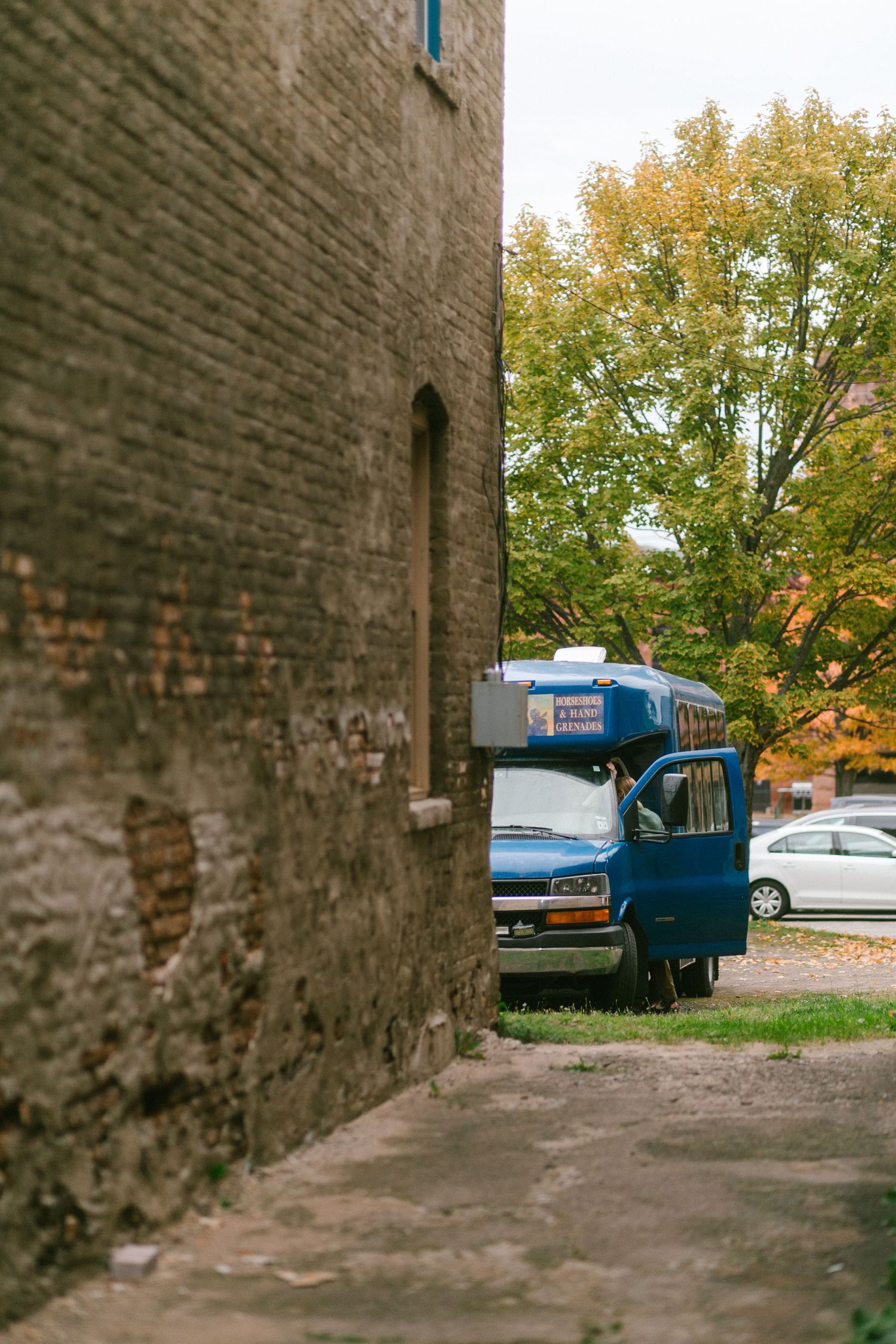 Old brick wall leads to parking area with vintage blue car and autumn trees in background.