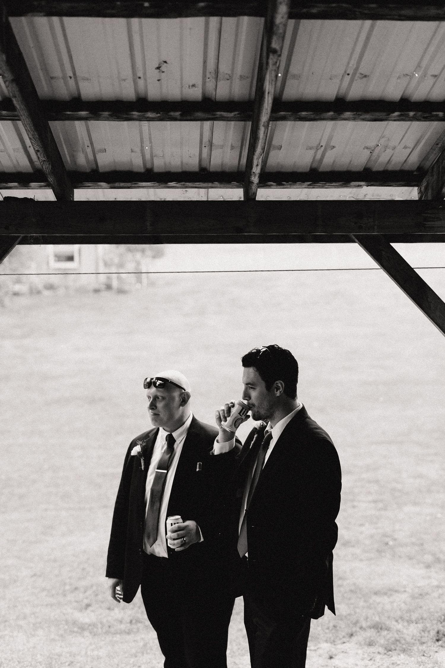 Two men in suits and sunglasses stand beneath a metal overhang, holding beverages in black and white photography.